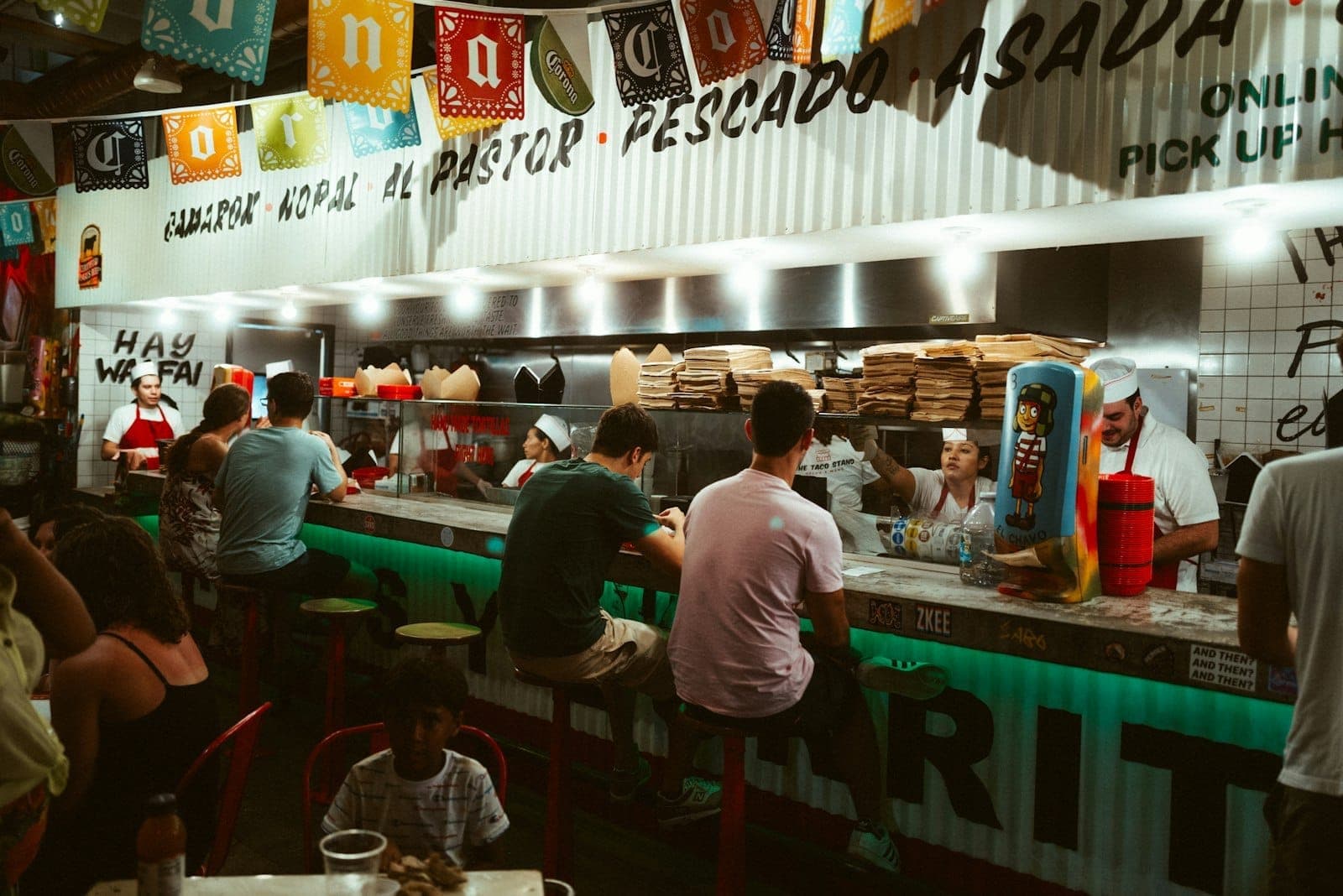 Des personnes assises dans un taco bar animé avec des cuisiniers préparant des plats derrière le comptoir, des décorations festives et des menus affichés au-dessus.