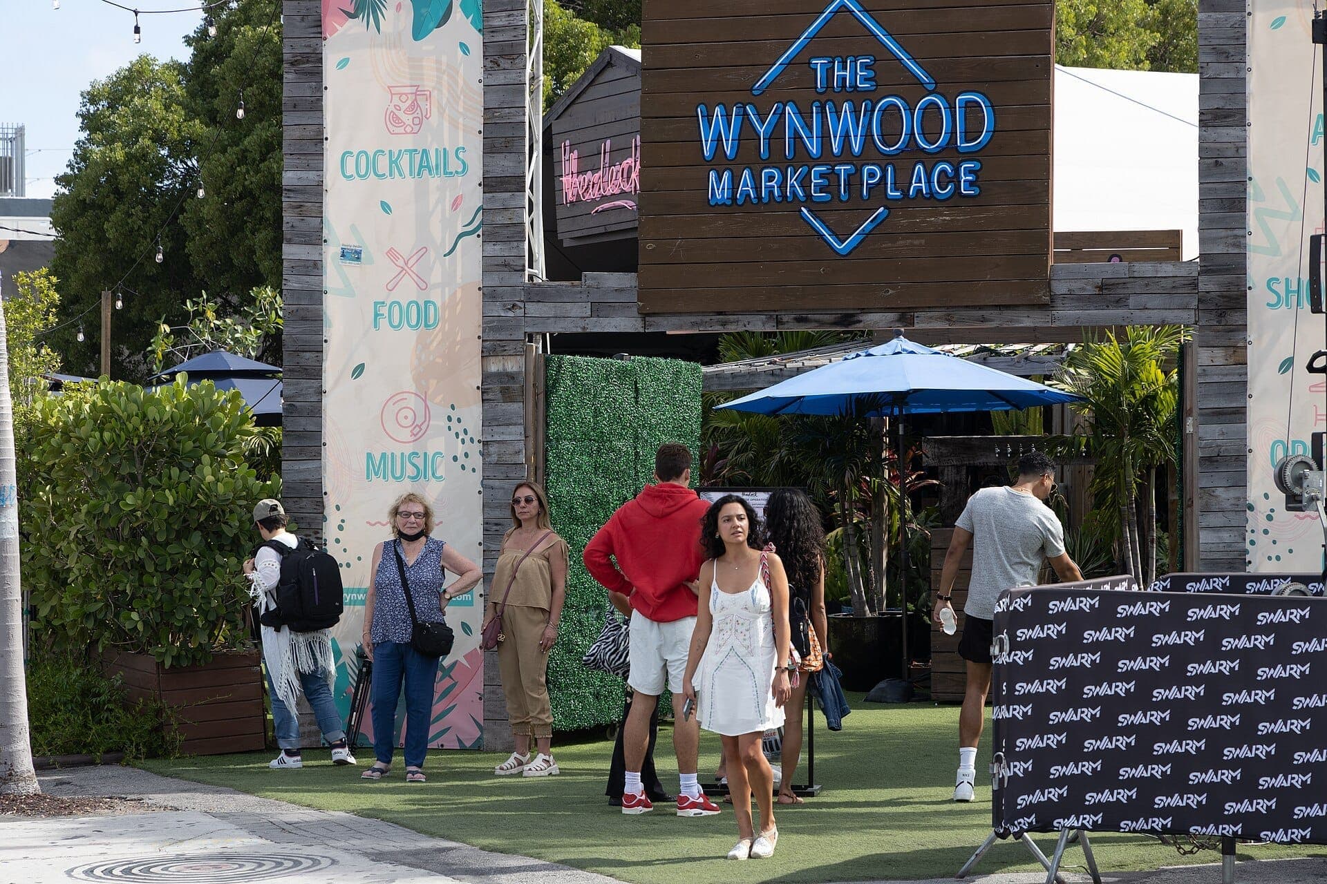 Entrée du Wynwood Marketplace à Miami avec des groupes de personnes habillées de façon décontractée, une signalétique colorée et une végétation tropicale en pleine lumière du jour.