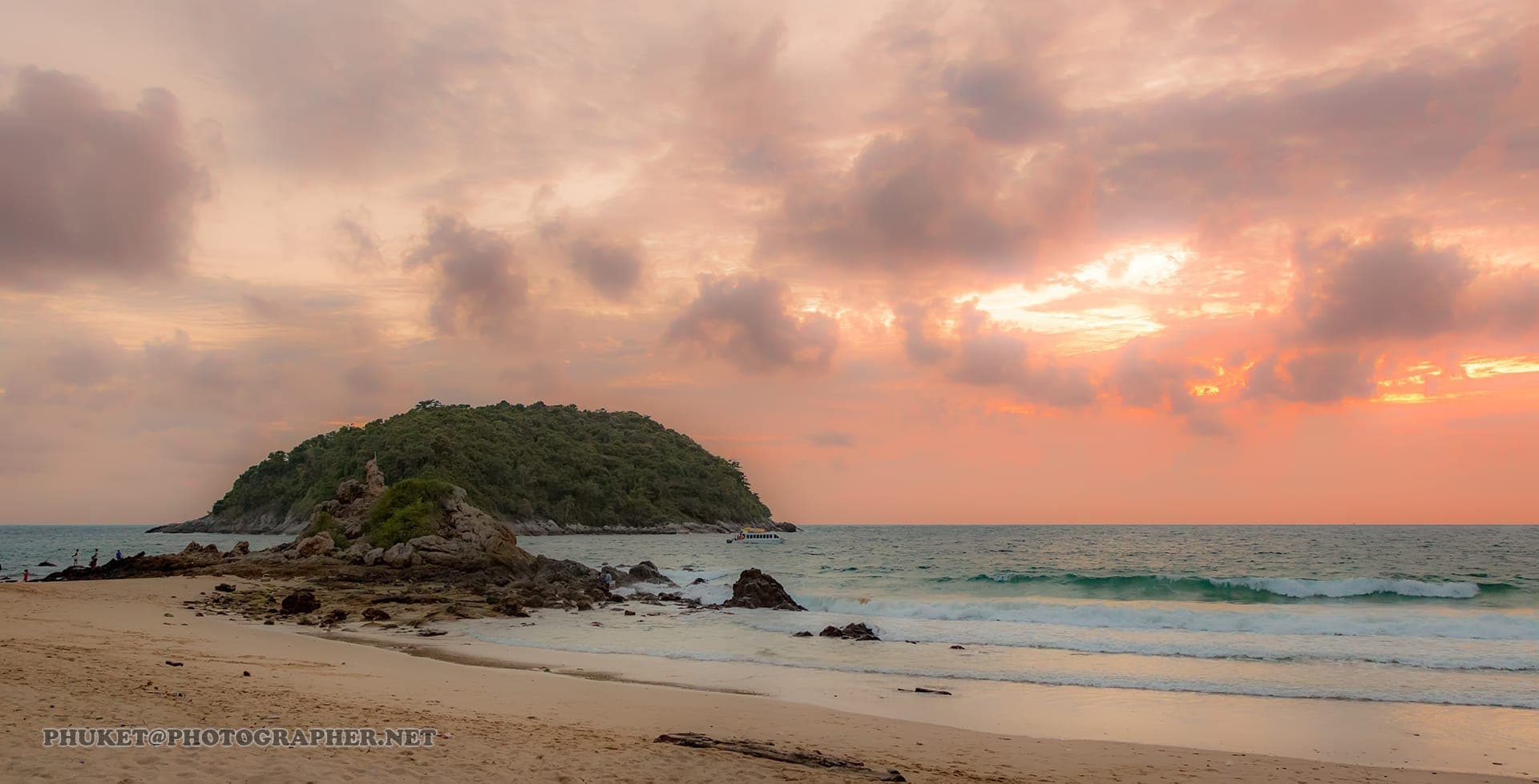 Wide sandy shore, rocky outcrops, and lush green hills at Ya Nui Beach in Phuket, with gentle waves under a pink-orange sunset sky.