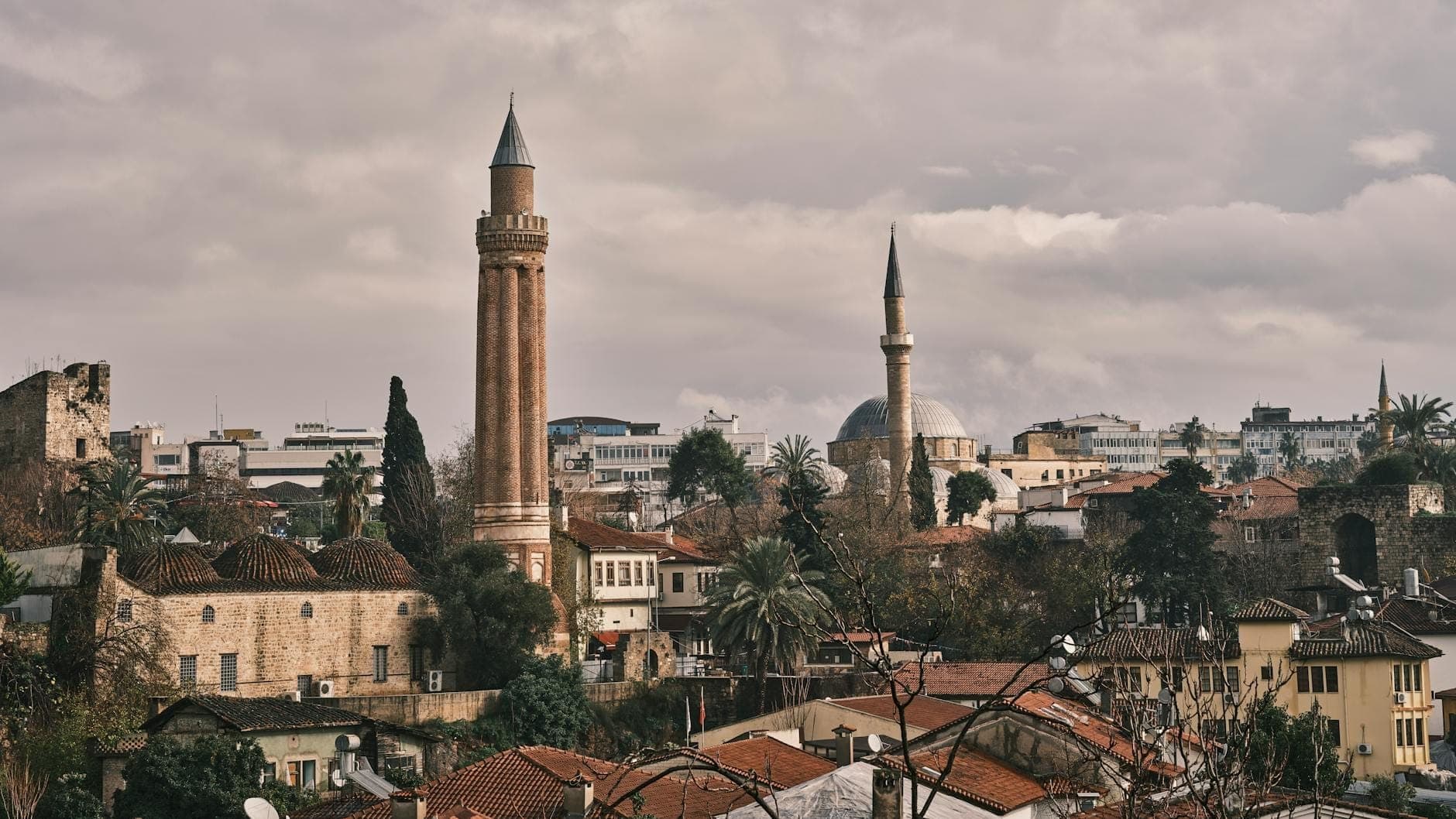 Panoramablick auf das Yivli Minare, das über den Dächern der Antalyaer Altstadt unter bewölktem Himmel aufragt, umgeben von historischen Gebäuden und üppigem Grün.