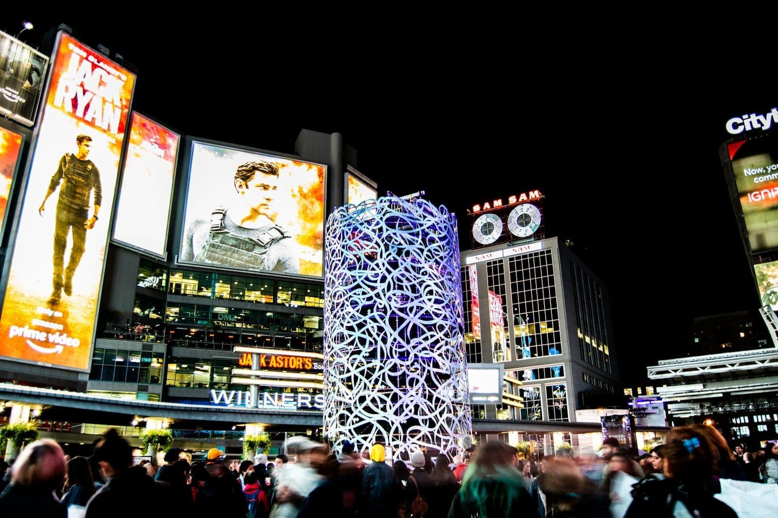 Sankofa Square (ex Yonge-Dundas Square) a Toronto di notte, con luci brillanti, grandi schermi digitali, folla di persone e un'energia urbana vivacissima.