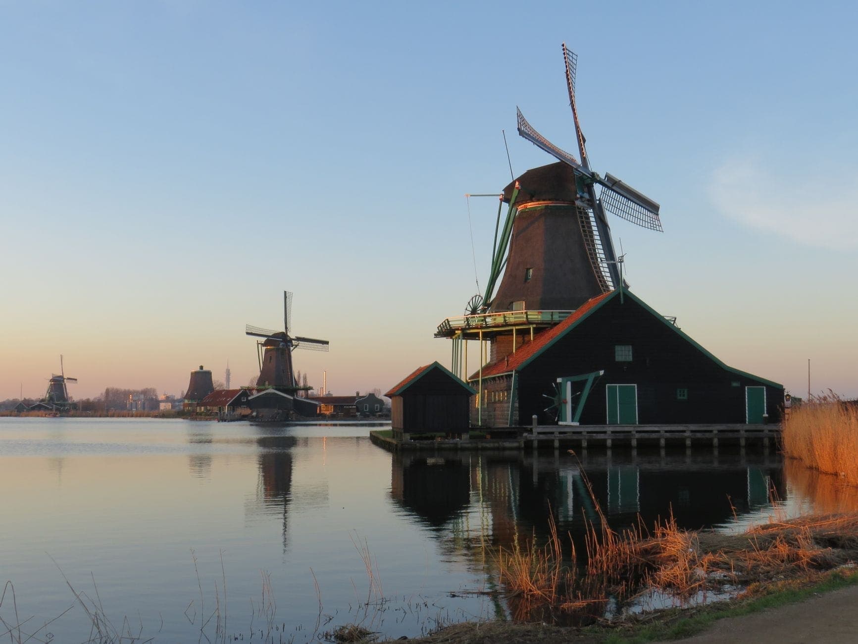 Klassische holländische Windmühlen und grüne Holzhäuser spiegeln sich im stillen Wasser von Zaanse Schans bei klarem Sonnenauf- oder -untergang.