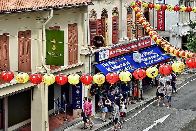 วิวยามค่ำของผู้คนต่อคิวหน้าแผงลอยอาหารสว่างไสว มีเนื้อย่างแขวนเรียงราย ในฮอว์กเกอร์เซ็นเตอร์ของสิงคโปร์