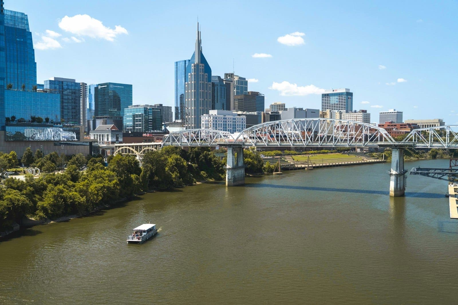 Wide view of Nashville skyline with the Batman Building, modern skyscrapers, pedestrian bridge, Cumberland River, and a boat cruising under a clear blue sky.