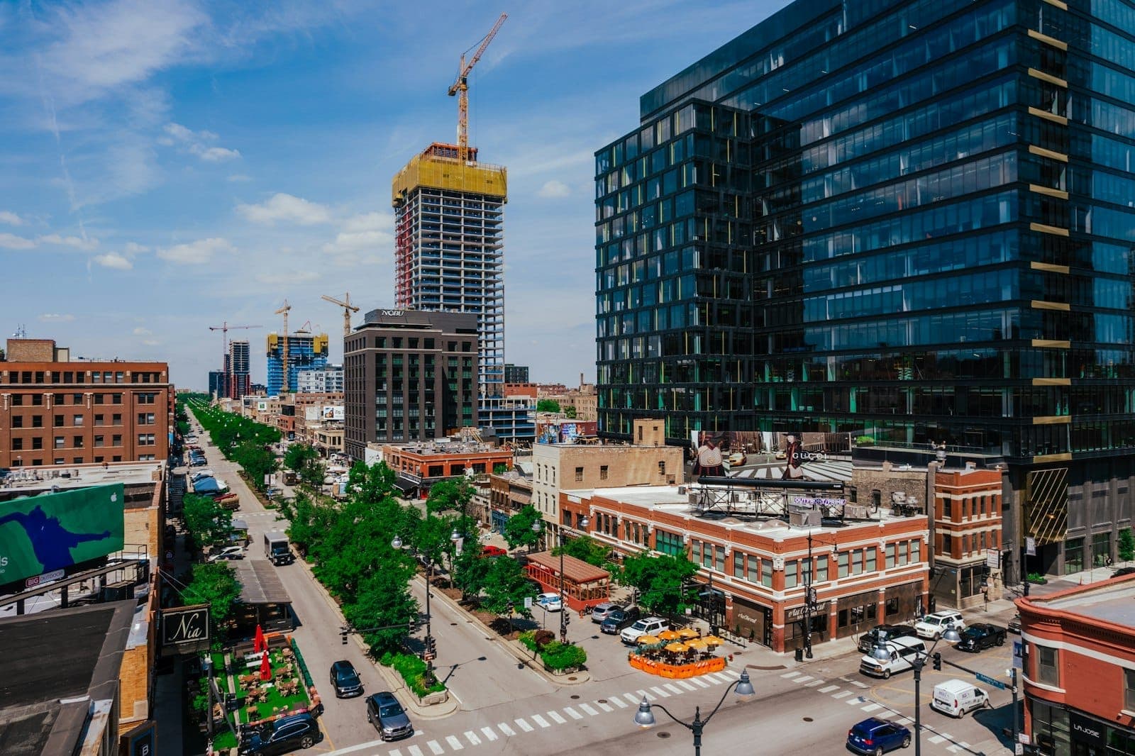 Animada calle urbana en un barrio de Chicago con nuevos edificios de gran altura, aceras arboladas, restaurantes y grúas al fondo.