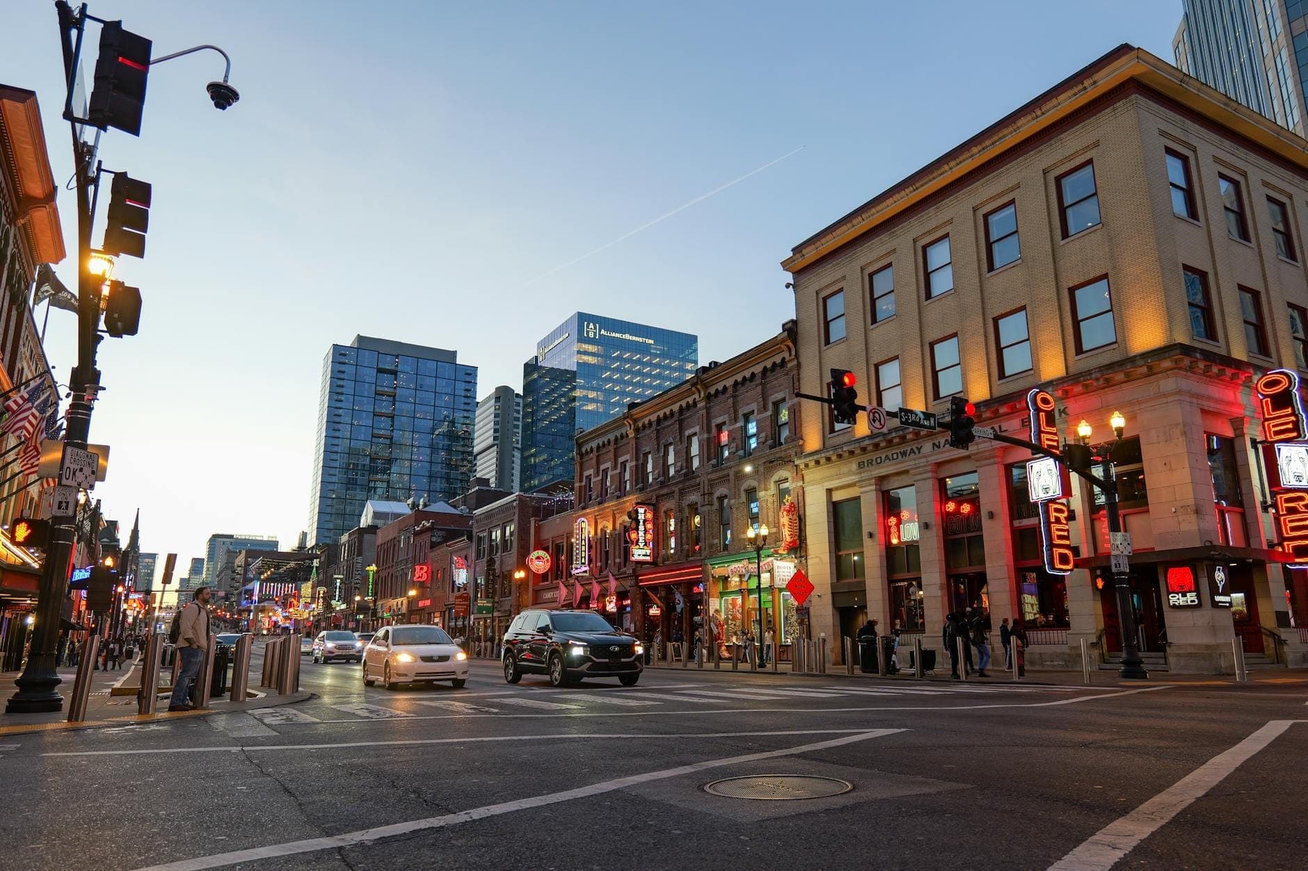 Tętniący życiem Lower Broadway w Nashville o zmierzchu, z neonowymi szyldami, barami i samochodami wzdłuż ożywionej ulicy oraz nowoczesnymi budynkami w tle.