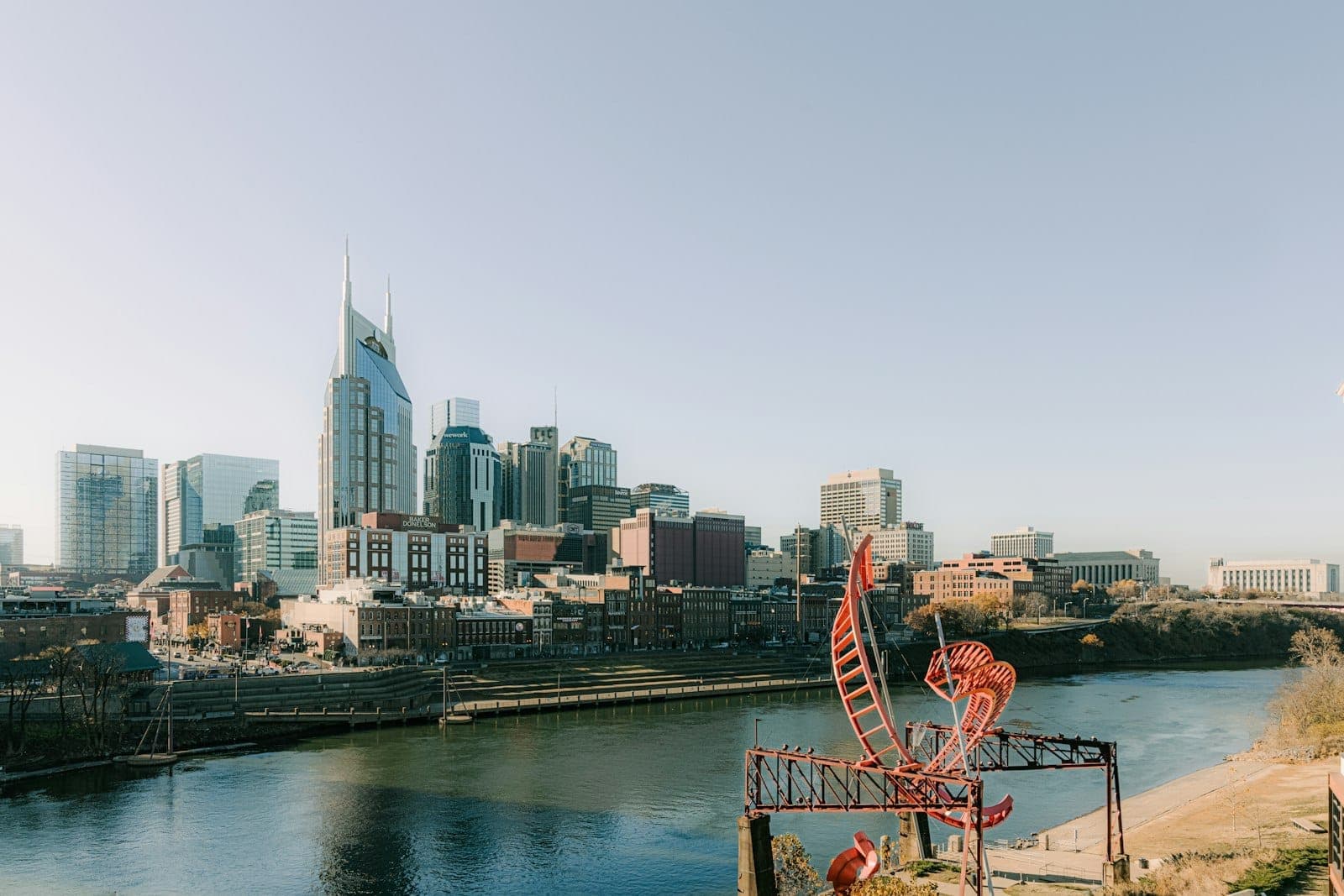 Wide view of Nashville skyline with river in foreground and iconic red sculpture, under clear blue sky on a sunny day.