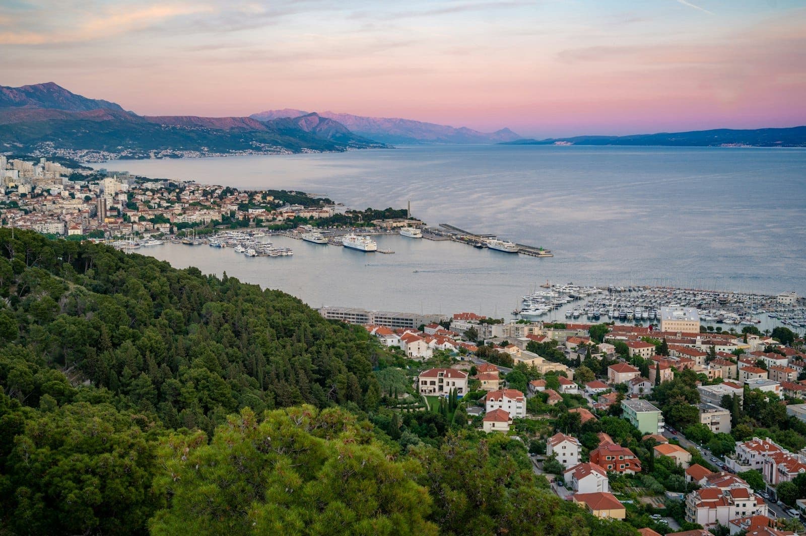 Vista panoramica della costa di Spalato con la collina Marjan, i quartieri della città, il porto turistico e le montagne lontane al tramonto.