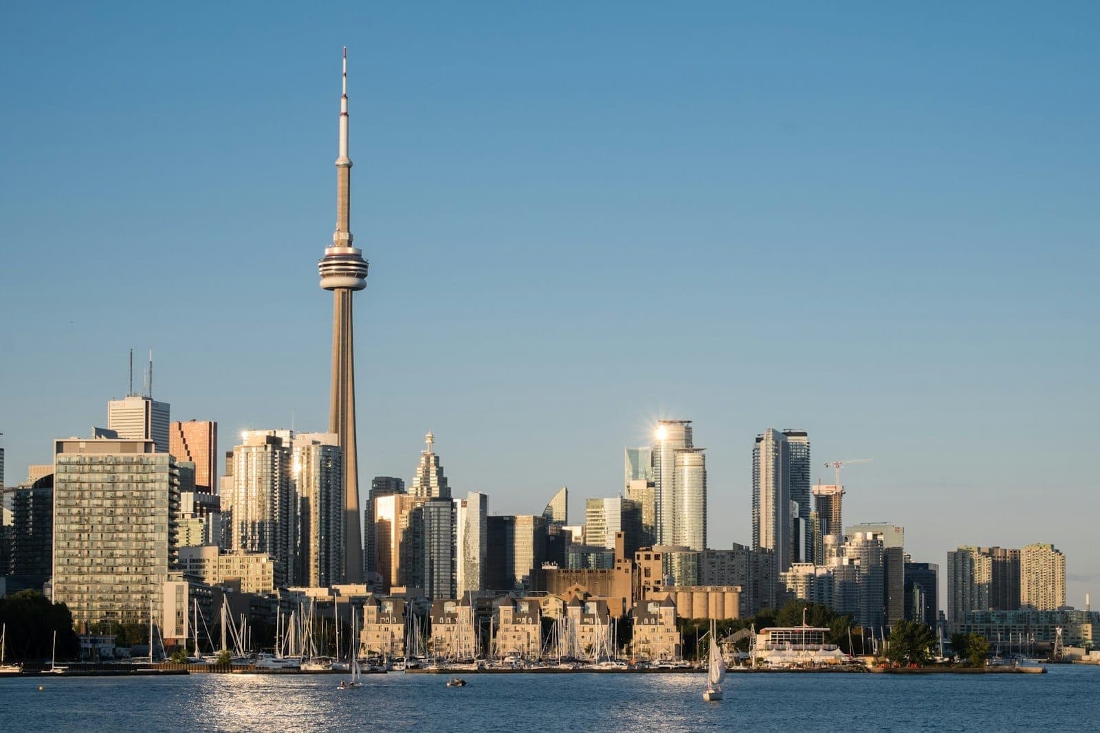 Vista diurna nitida dello skyline di Toronto dal lungomare con la CN Tower e gli edifici, barche a vela ormeggiate nel porto e cielo blu.