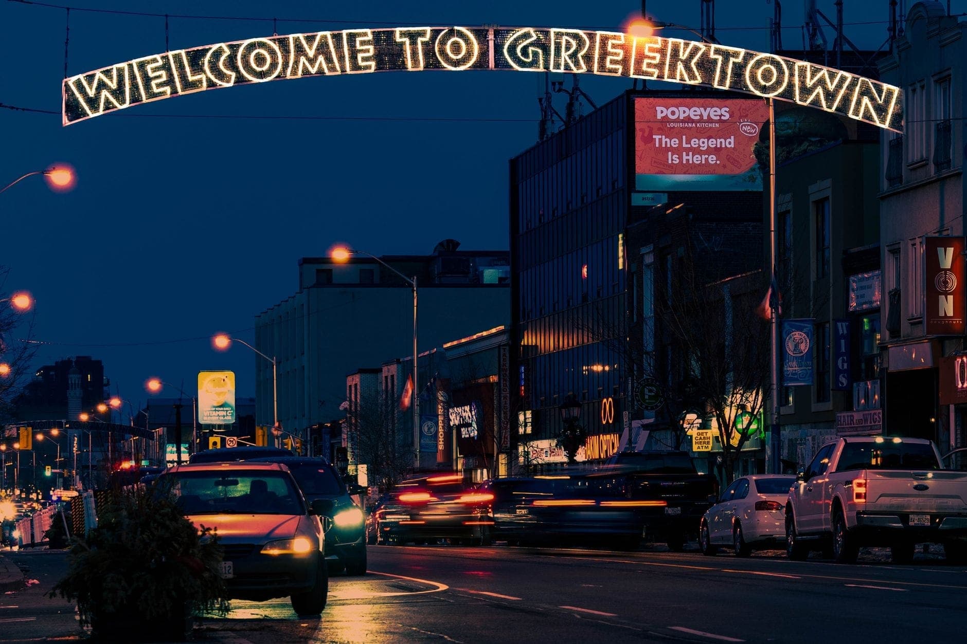 Scena notturna in Greektown a Toronto con un luminoso cartello 'Welcome to Greektown' e auto sulla strada.