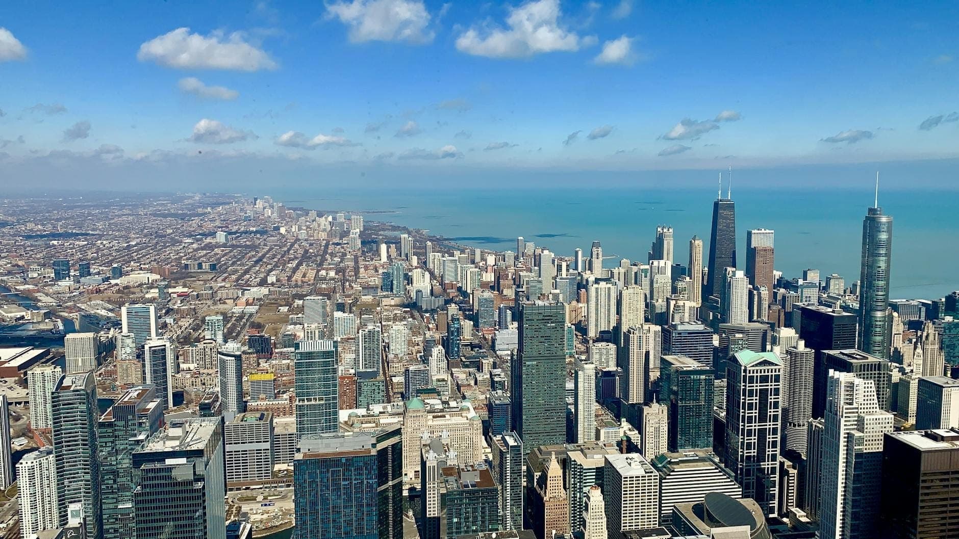 Wide panoramic view of Chicago’s skyline and Lake Michigan on a sunny day, as seen from a high observation deck with blue sky and scattered clouds.