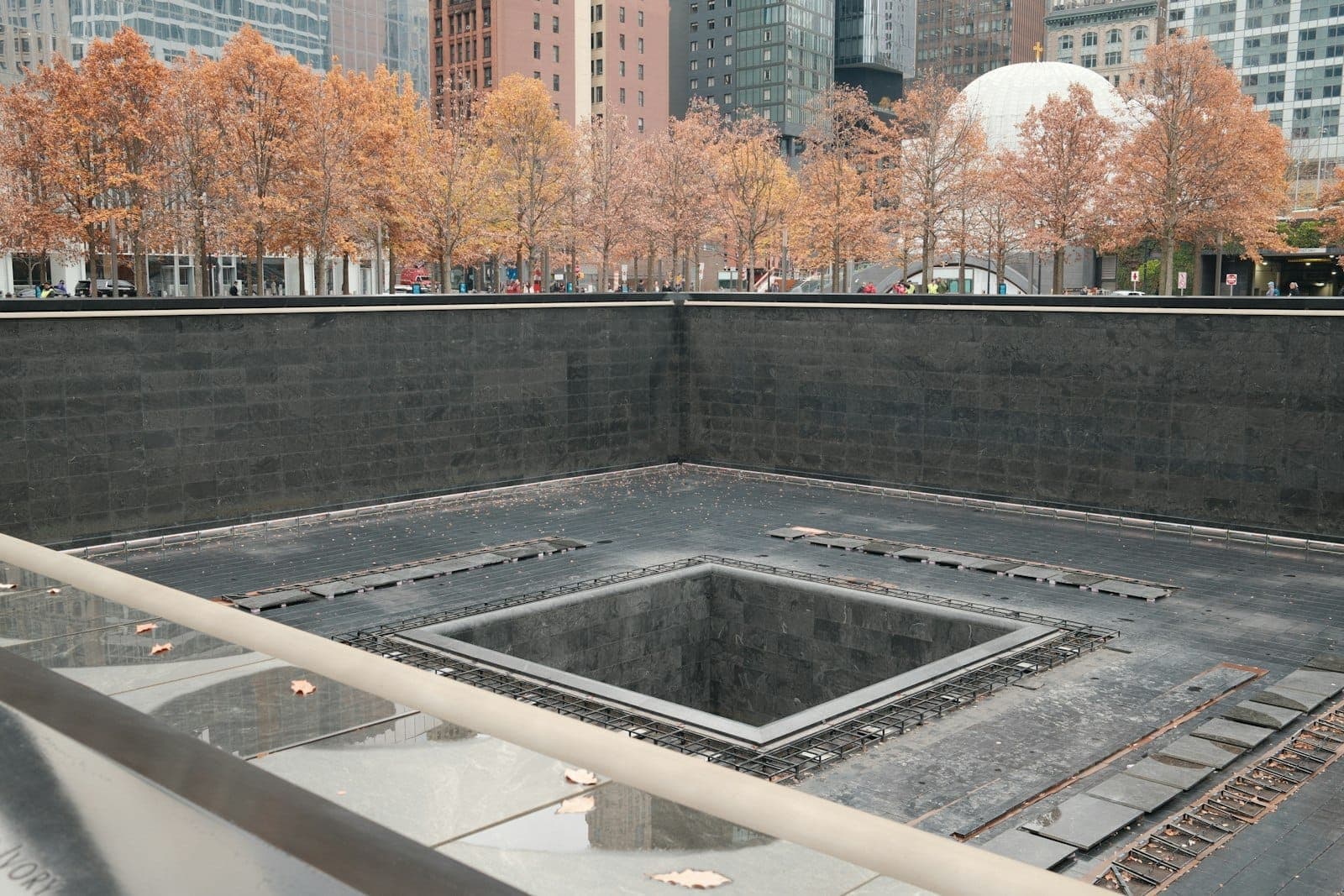 สระสะท้อนน้ำของ National September 11 Memorial พร้อมต้นไม้ในฤดูใบไม้ร่วงและอาคารในเมืองเป็นฉากหลังในวันที่มีเมฆมาก