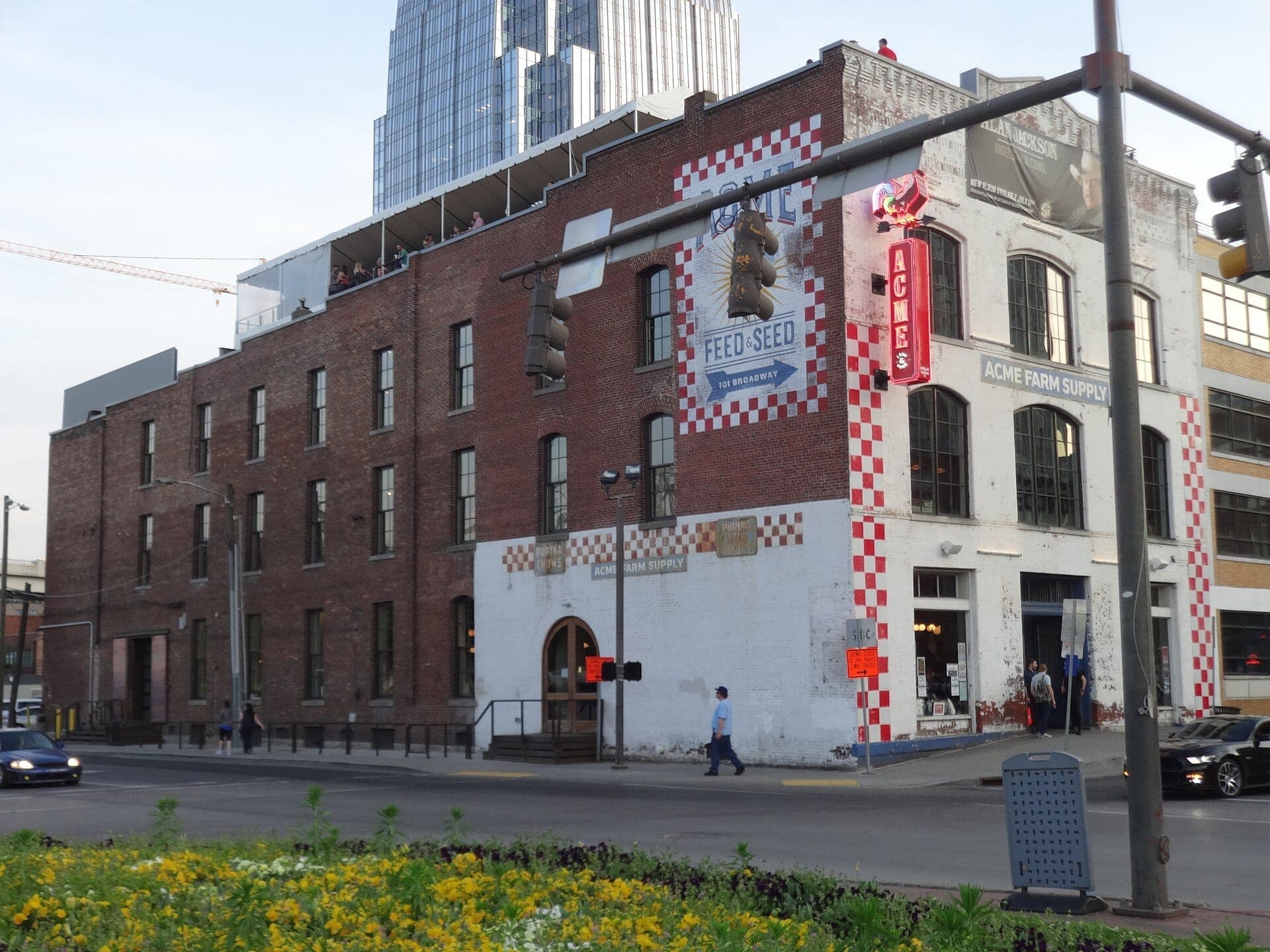 Exterior view of Acme Feed & Seed, a historic brick building on Lower Broadway with red accents, neon sign, and rooftop patrons at dusk.