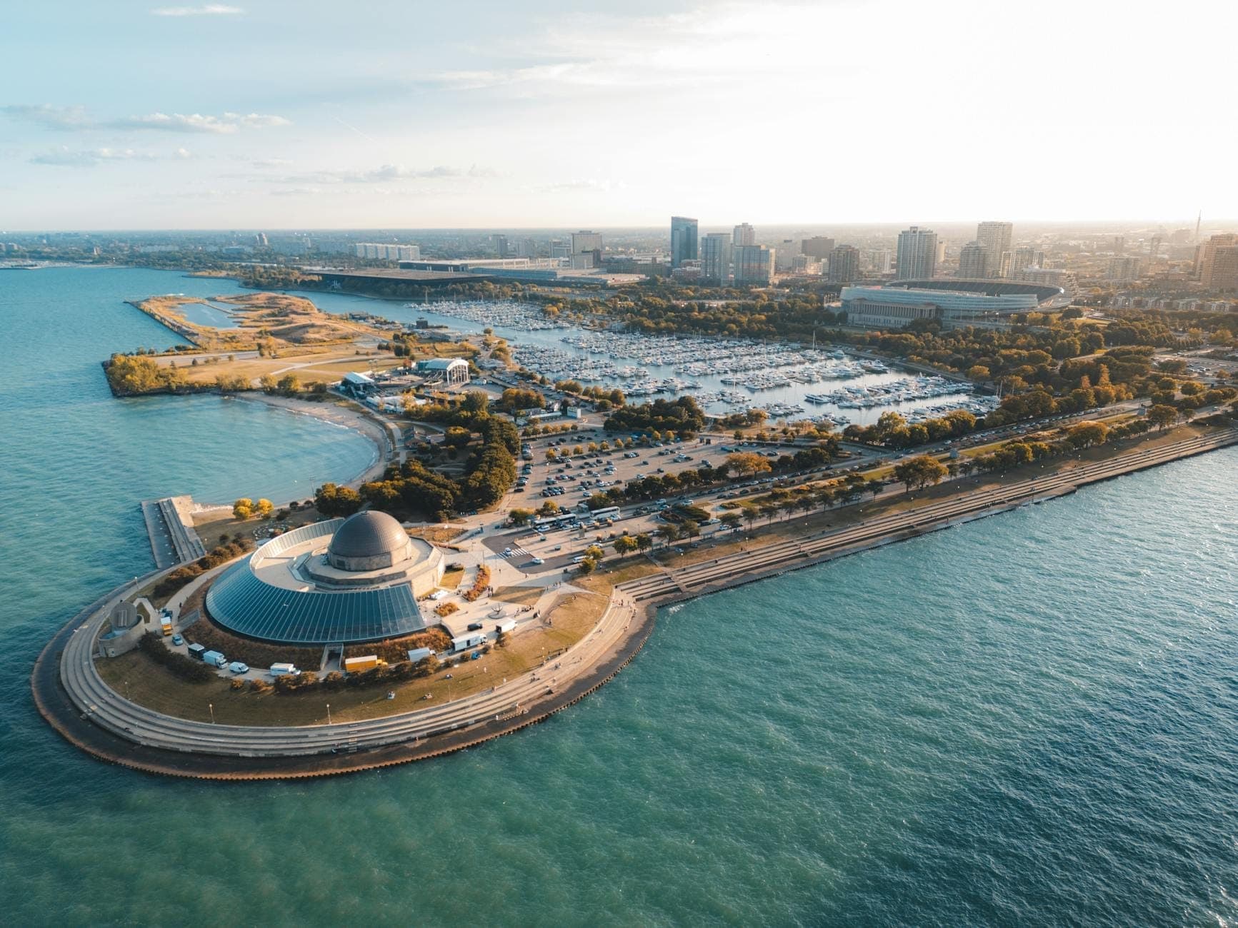 Aerial view of Adler Planetarium at the tip of a peninsula, surrounded by Lake Michigan with the Chicago skyline in the background on a sunny day.