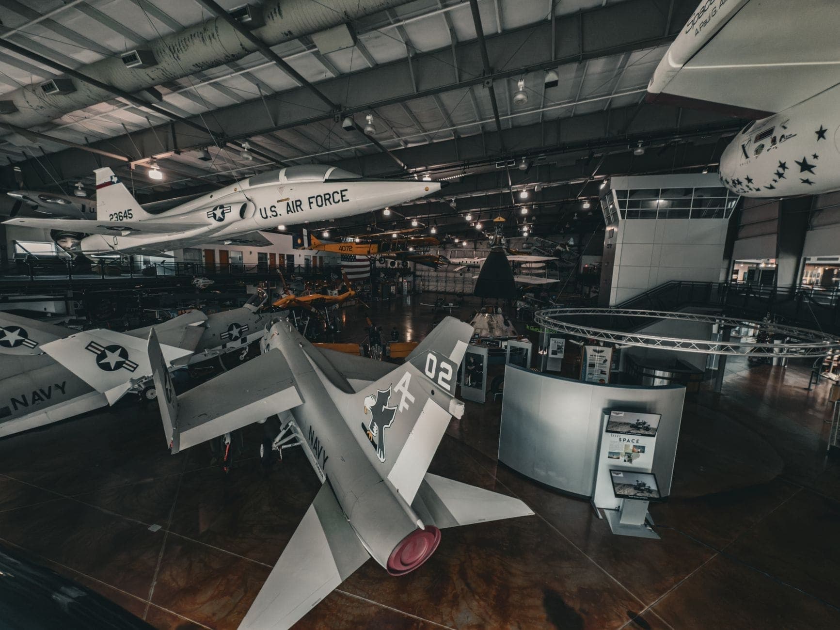 Interior view of an aviation museum exhibit with various historic airplanes, including a US Air Force jet, under a large metal roof.