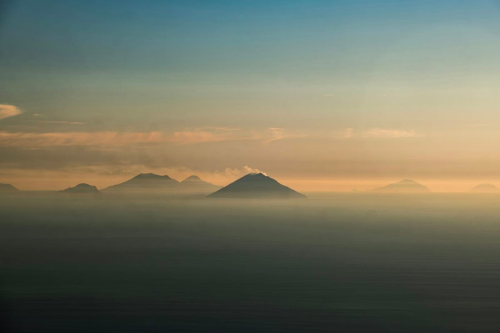 Pemandangan dramatis beberapa Kepulauan Aeolian dengan puncak-puncak vulkanik yang menjulang dari lautan berkabut saat matahari terbenam, menciptakan suasana perjalanan yang indah dan mengundang.