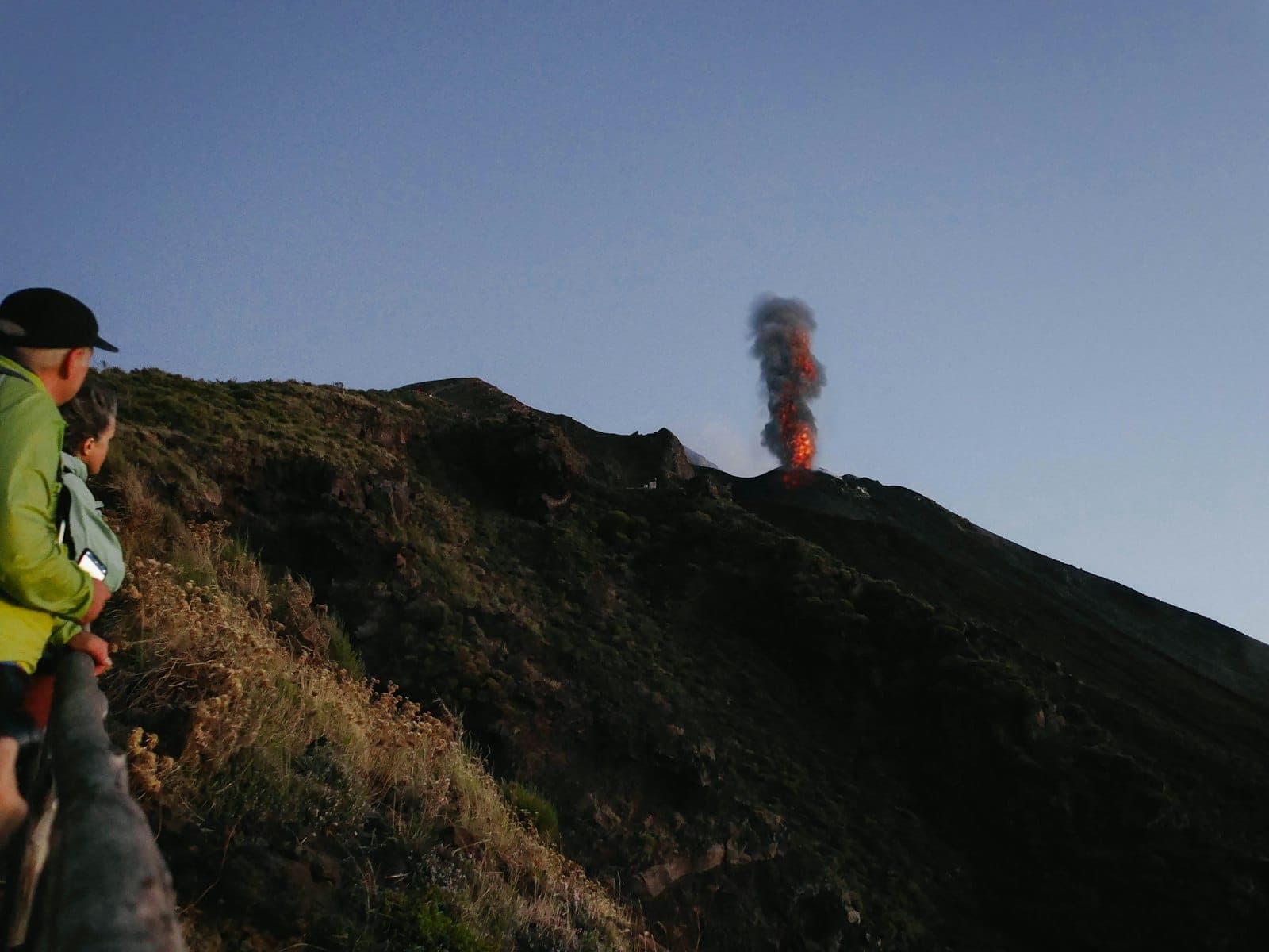 Dua orang menyaksikan letusan gunung berapi Stromboli saat senja di lereng berbatu yang hijau di Kepulauan Aeolian, dengan asap dan lava yang terlihat.