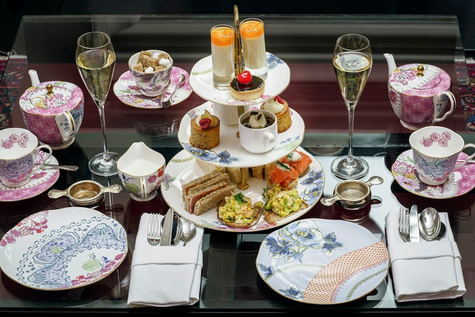 Elegante servizio di tè pomeridiano con vassoio a piani di tramezzini, pasticcini e scones, tazze da tè floreali e calici di champagne su un tavolo.