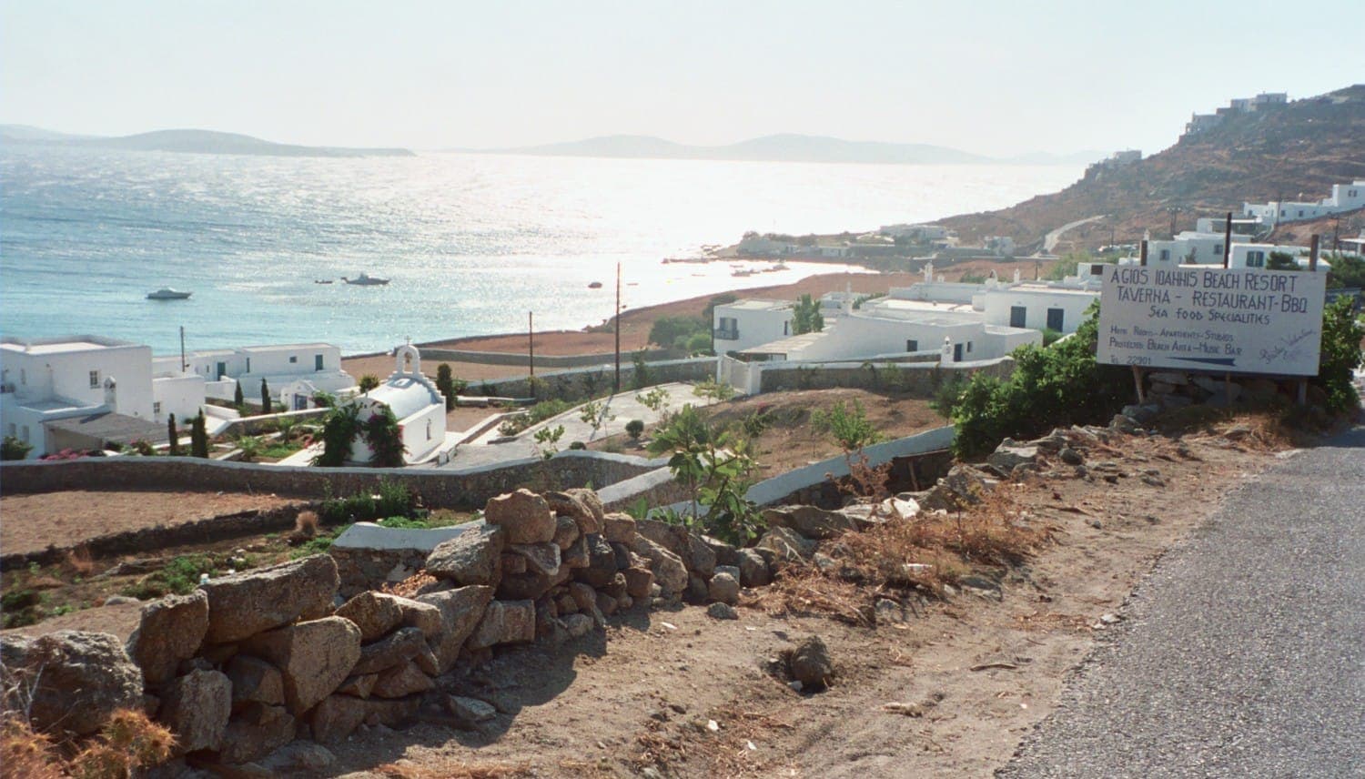 Blick auf den Strand Agios Ioannis in Mykonos, mit weiß getünchten Gebäuden, einer felsigen Küste und türkisfarbenem Meer unter der hellen Nachmittagssonne.