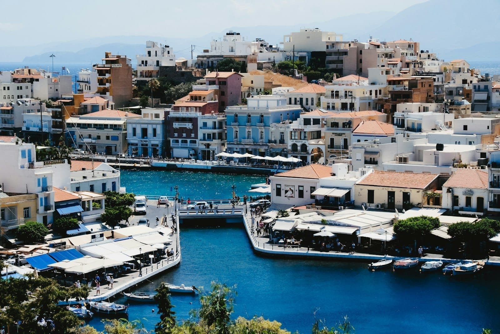 Eine lebhafte Küstenstadt rund um einen blauen Salzsee mit Booten, Cafés am Wasser und bunten Häusern, die sich die Hügel hinaufziehen – Agios Nikolaos auf Kreta.