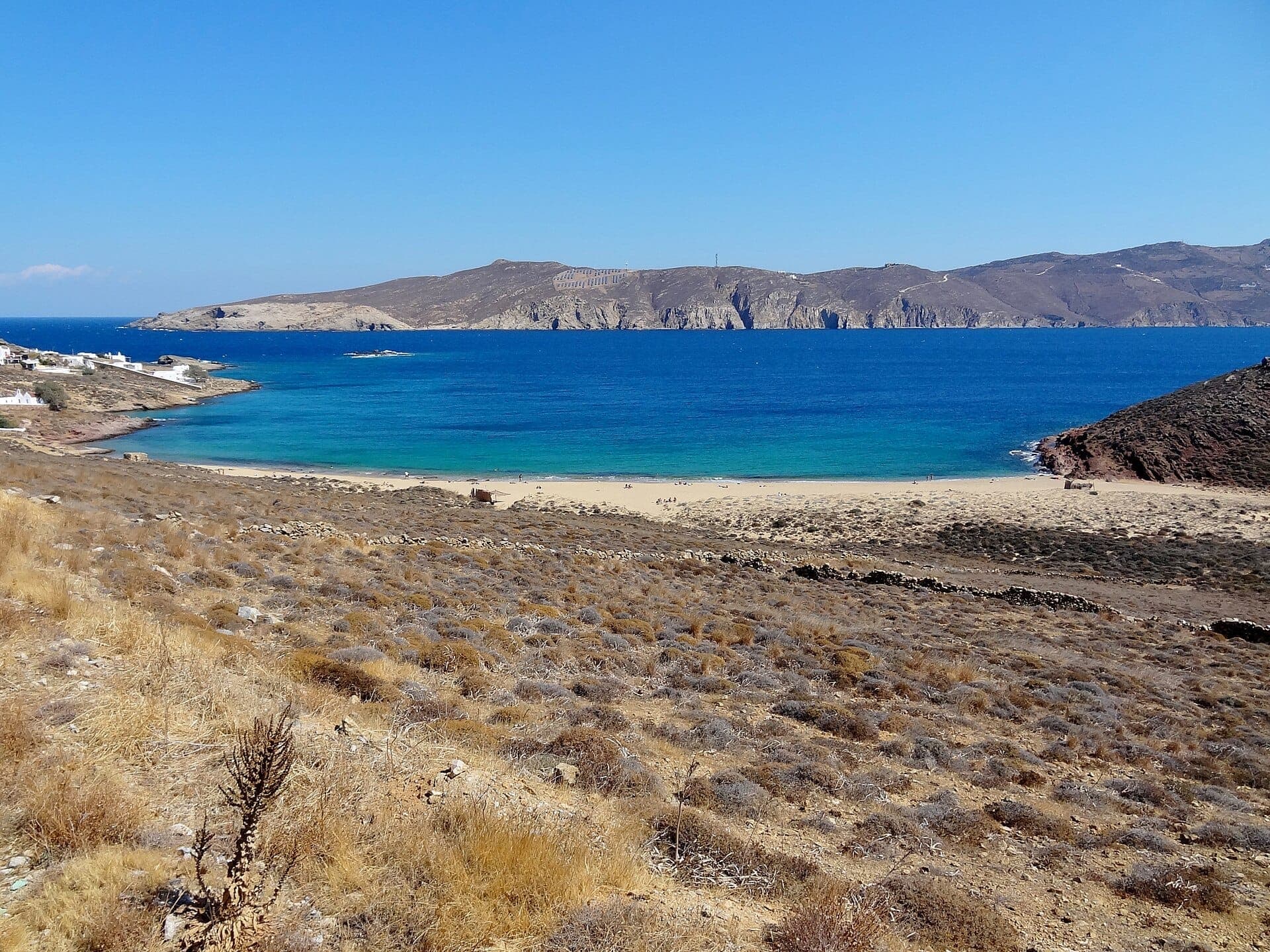 Weitläufige Ansicht des Agios Sostis Beach auf Mykonos mit goldenem Sand, klarem blauem Meer, trockenen Hügeln und verstreuten weiß getünchten Gebäuden.