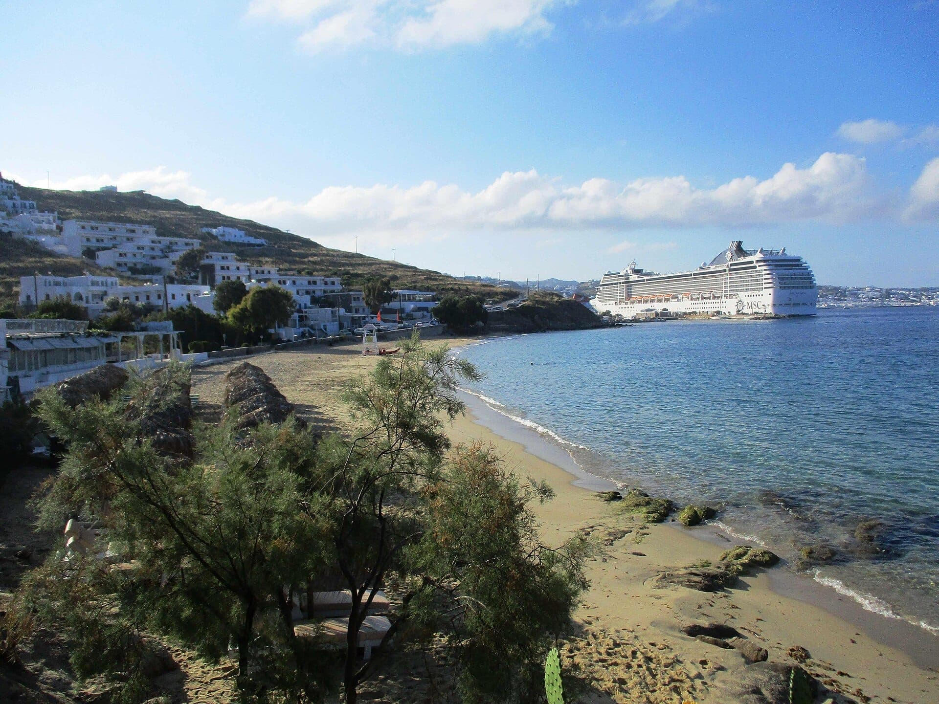 Agios Stefanos Beach mit ruhigem Sandstrand, weiß getünchten Gebäuden am Hang und einem großen Kreuzfahrtschiff, das unter blauem Himmel nahe der Küste vor Anker liegt.