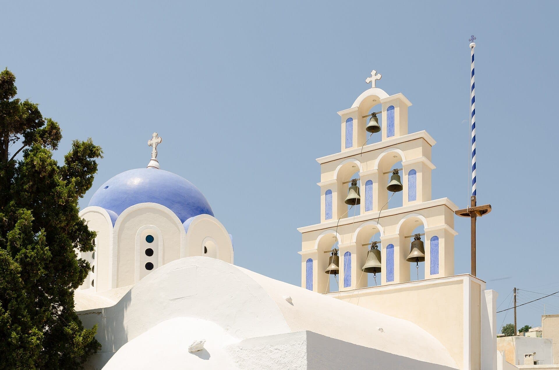 Weiß getünchte Kirche mit blauer Kuppel und traditionellem Glockenturm in Akrotiri, Santorini, unter klarem blauem Himmel mit hellem Sonnenlicht.