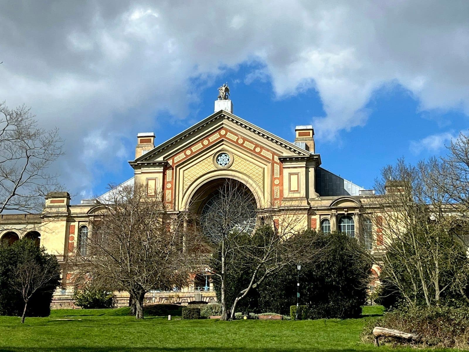 Fasad Victoria yang megah dari Alexandra Palace menjulang di atas taman hijau, dikelilingi pepohonan dengan latar langit biru cerah dan awan yang dramatis.
