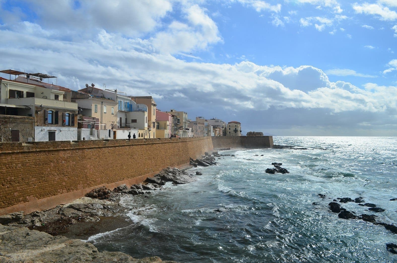 Vista de las murallas medievales de Alghero frente al mar, con edificios históricos que se asoman al Mediterráneo bajo un cielo parcialmente nublado y olas rompiendo contra la orilla rocosa.