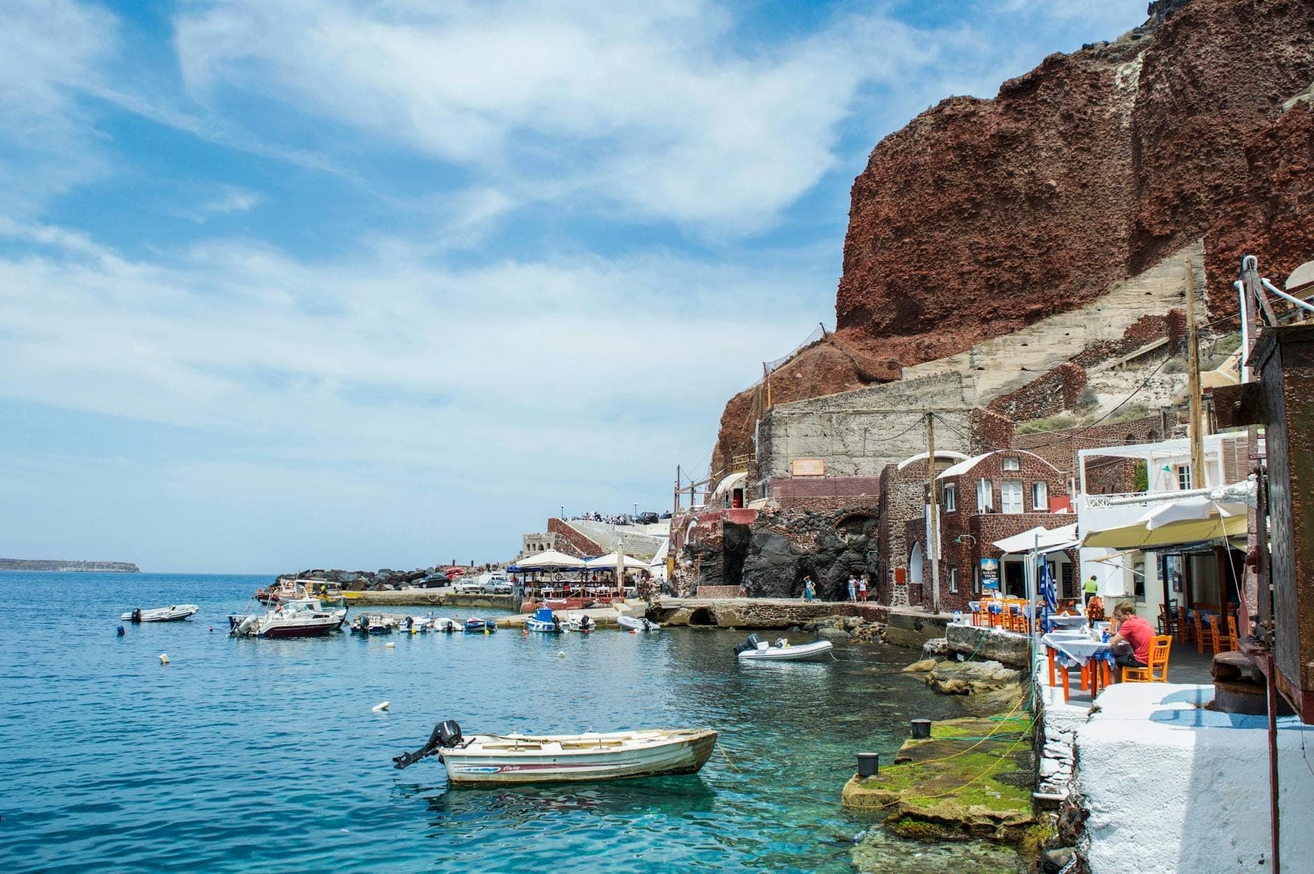 Die Ammoudi Bay mit türkisfarbenem Wasser, festgemachten Booten, Fischrestaurants direkt am Wasser und dramatischen roten Vulkanfelsen dahinter unter einem teils bewölkten Himmel.