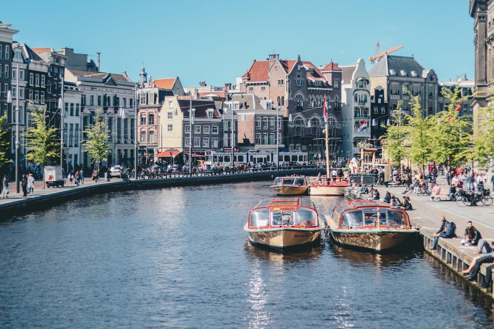 Amsterdamer Kanal mit historischen Gebäuden, angedockten Kanalkreuzfahrtbooten und Menschen, die unter einem klaren blauen Himmel entlang der Uferpromenade sitzen.