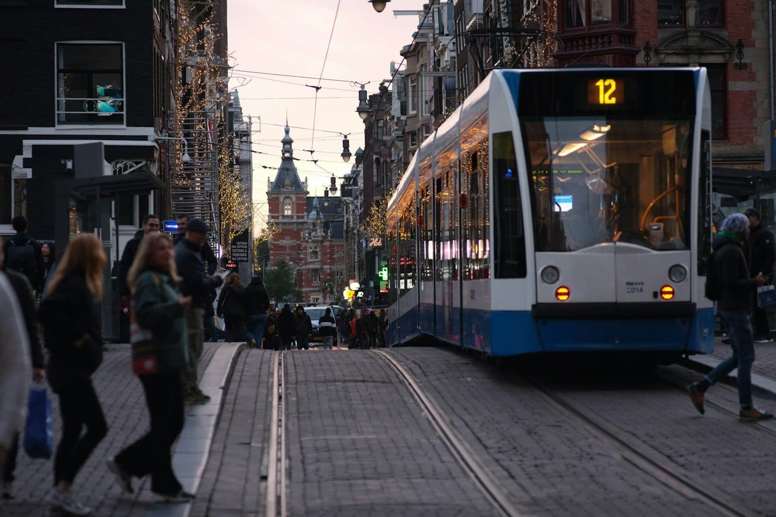 Eine blau-weiße Straßenbahn fährt durch eine belebte Straße in Amsterdam mit Fußgängern und traditionellen niederländischen Gebäuden im Hintergrund.