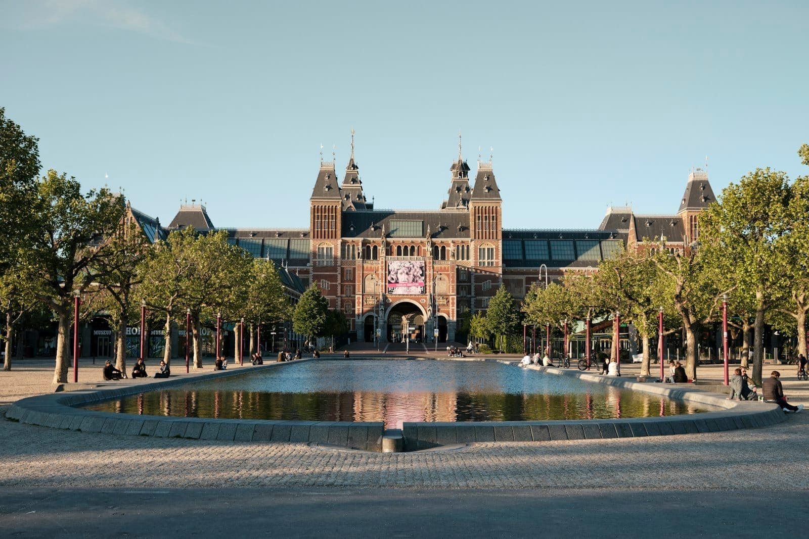 Weitblick auf das Rijksmuseum in Amsterdam mit Spiegelbecken und Bäumen auf dem Museumplein im Morgenlicht.