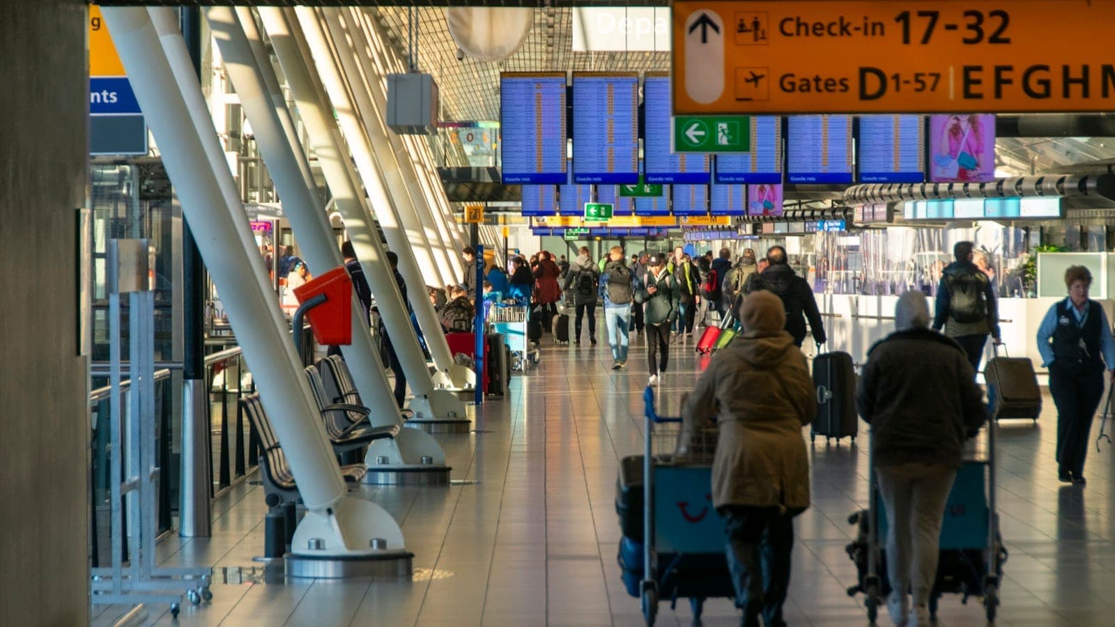 Belebtes Terminal am Flughafen Amsterdam Schiphol mit Reisenden, Gepäckwagen, Abflugtafeln und gelber Beschilderung unter heller Innenbeleuchtung.