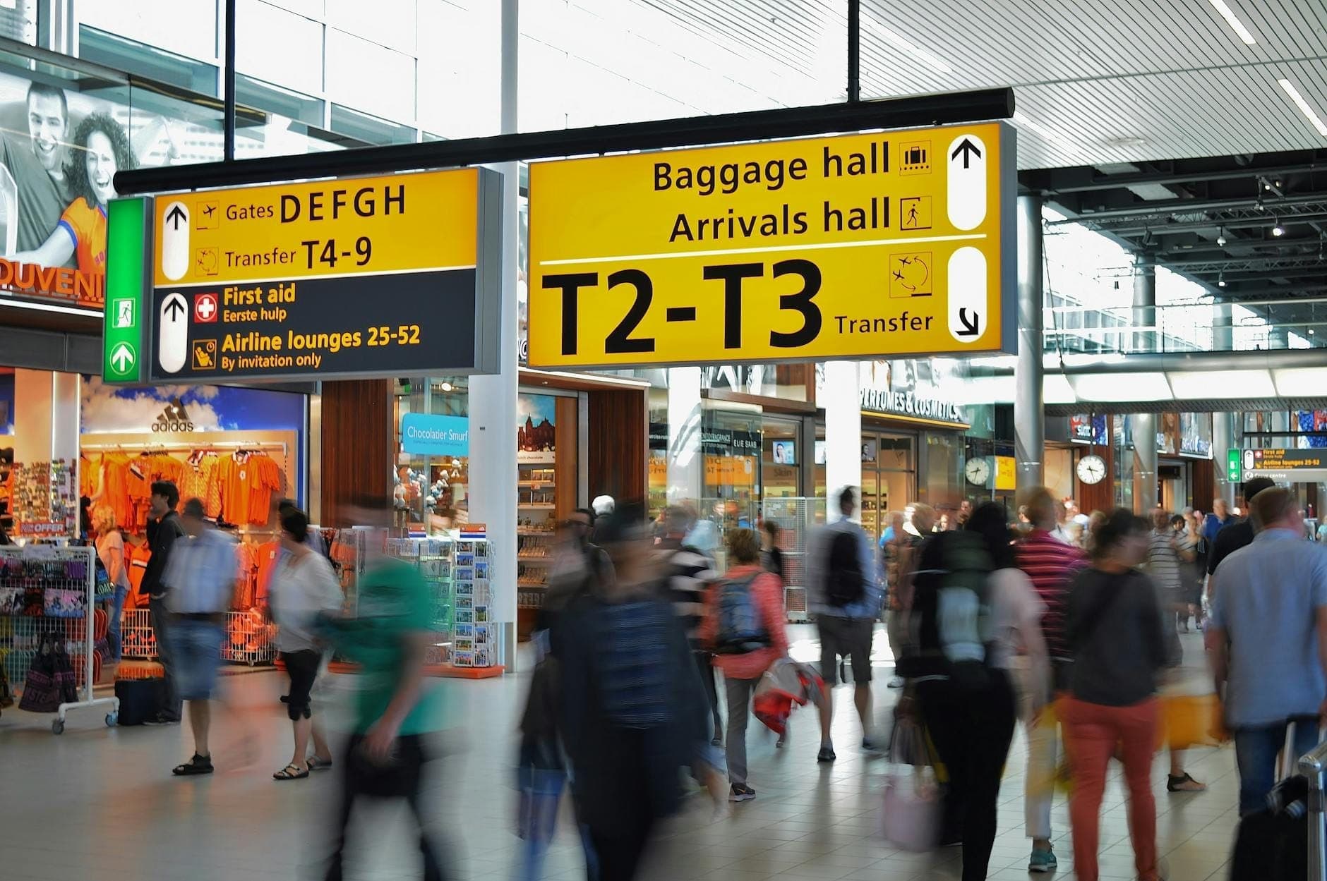 Belebte Innenszene am Flughafen Schiphol mit Geschäften, Cafés und Menschen, die unter hellen gelben Terminalschildern entlanggehen.