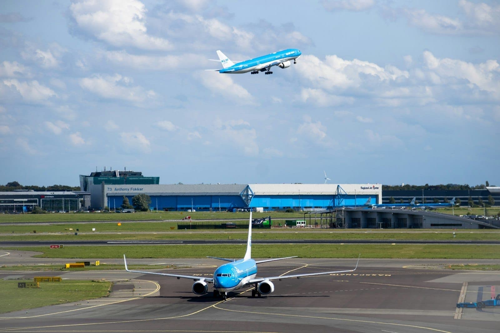 KLM-Flugzeuge auf der Start- und Landebahn und beim Abheben am Flughafen Amsterdam Schiphol, mit blauem Himmel und Flughafengebäuden im Hintergrund.
