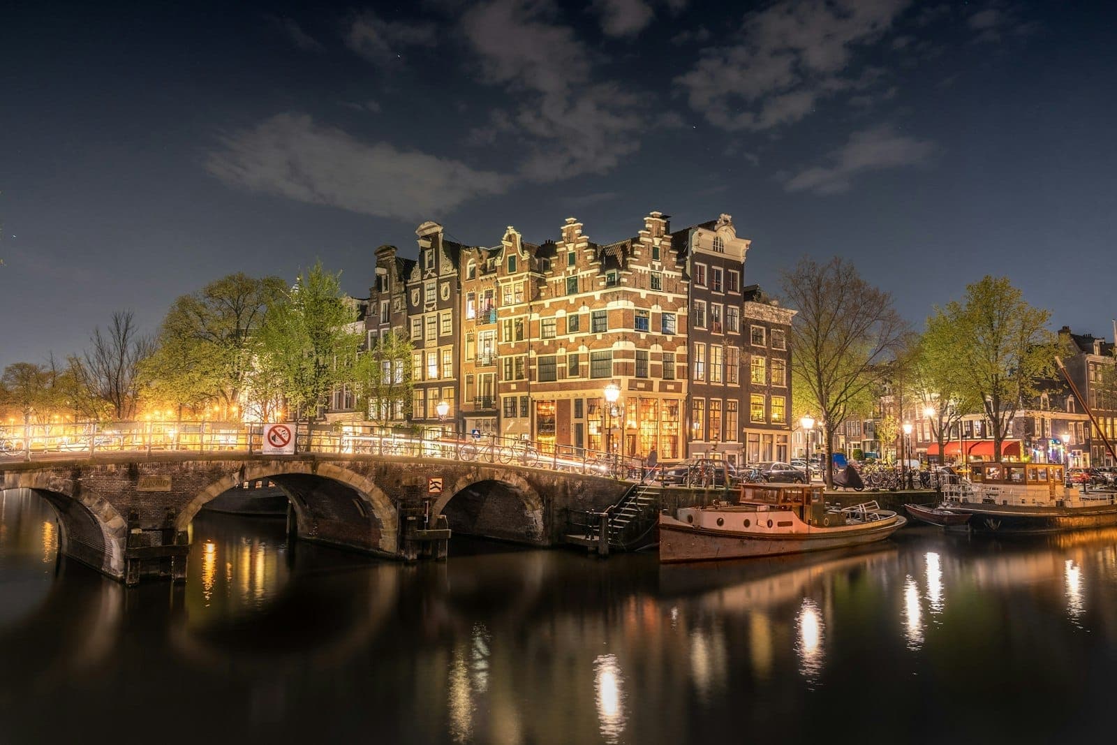 Nächtlicher Blick auf beleuchtete Amsterdamer Grachtenhäuser und eine Bogenbrücke, die sich im Wasser spiegeln, mit Booten und Bäumen unter dramatischem Himmel.