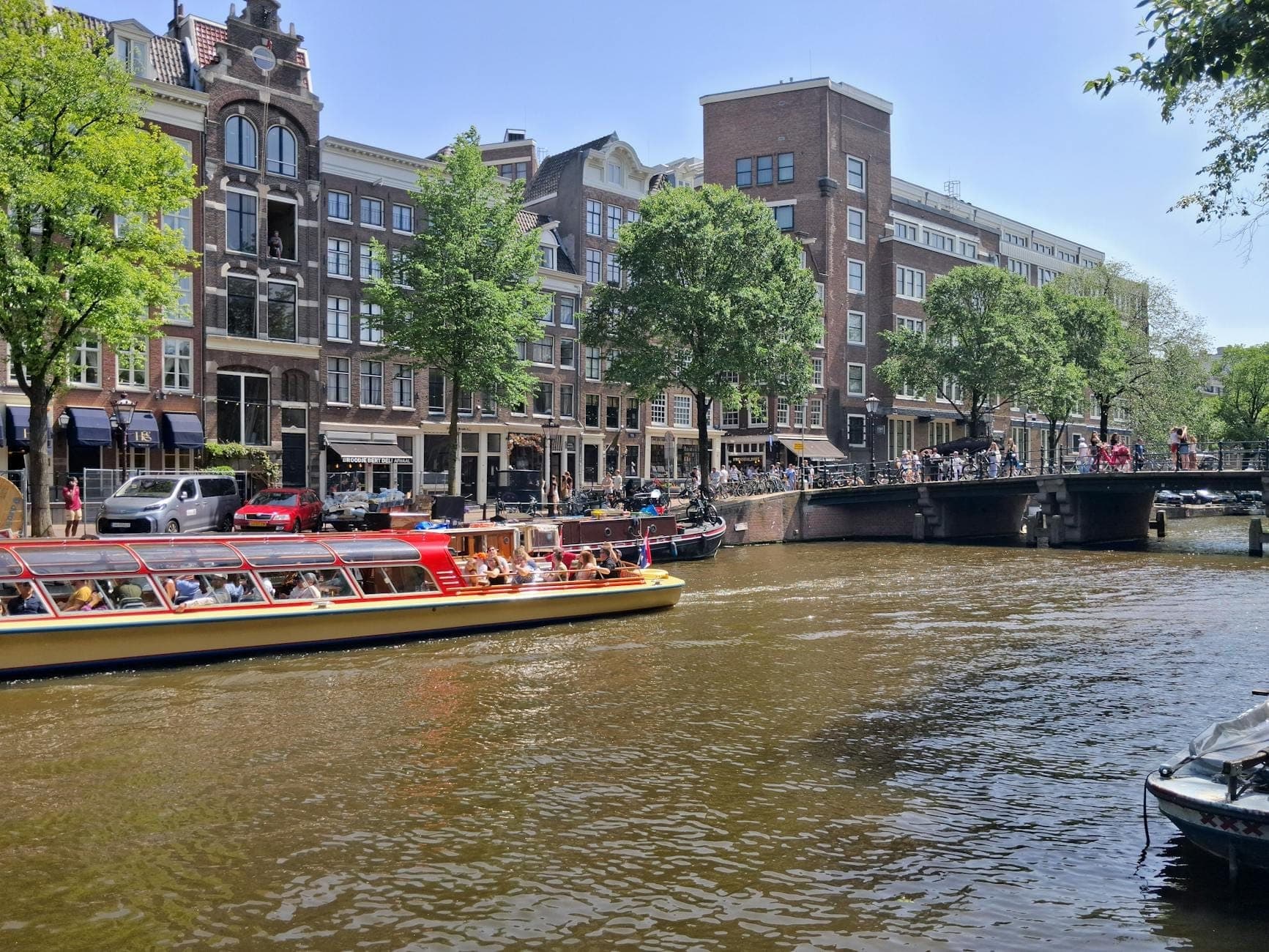 Sonnige Kanalszene in Amsterdam mit Kanalkreuzfahrtboot, klassischen niederländischen Häusern und Menschen, die das warme Wetter auf der Brücke genießen.