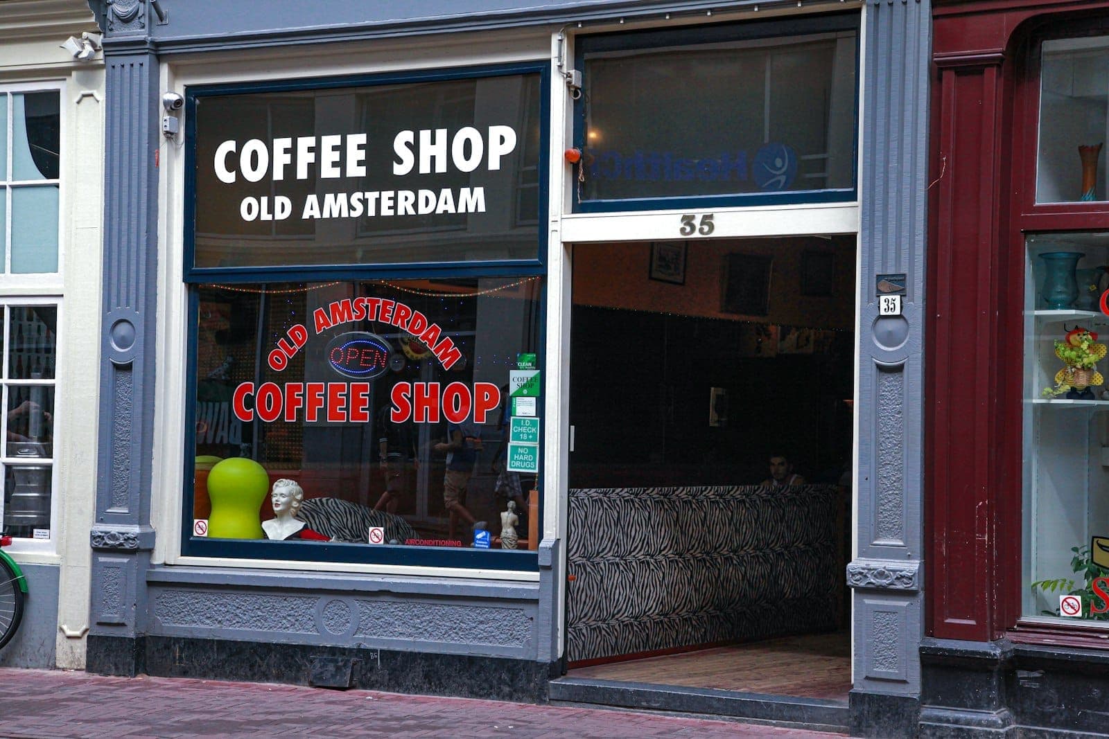 Der Eingang zum Coffee Shop 'Old Amsterdam' mit großen Fenstern, Beschilderung und einem Blick ins Innere mit Sitzgelegenheiten und typischer Einrichtung der Amsterdamer Coffeeshops.
