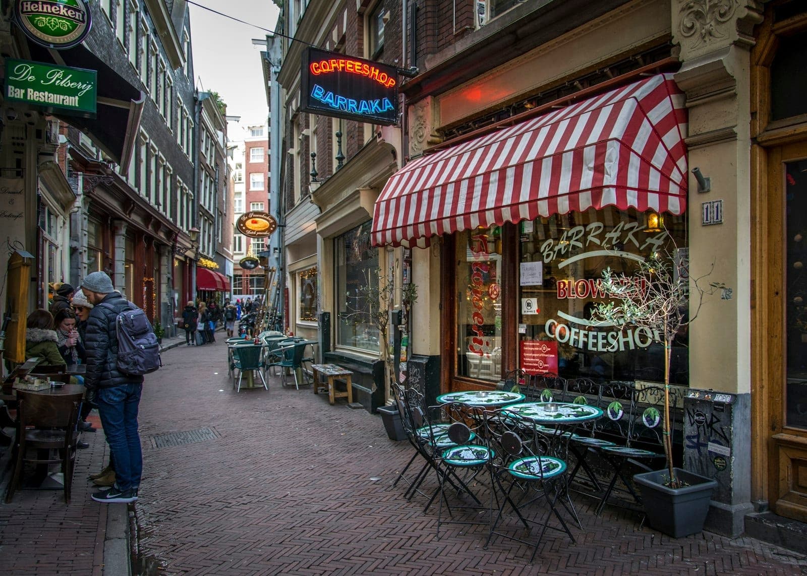 Straßenansicht eines klassischen Amsterdamer Coffeeshops mit Außentischen, Leuchtreklamen und Passanten in einer lebhaften, engen Gasse.