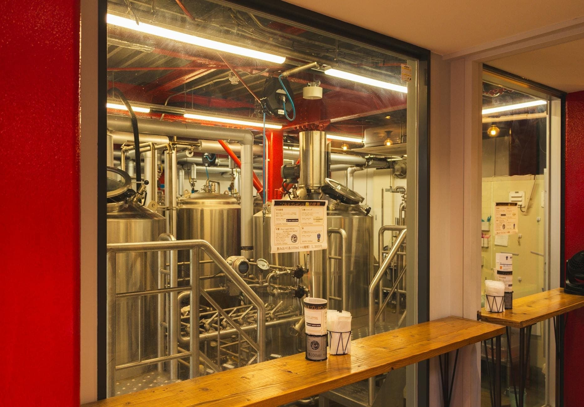 Innenansicht einer Craft-Brauerei mit hinter Glas sichtbaren Brauanlagen, in einem modernen Industrieraum mit roten Wänden und Thekensitzen.