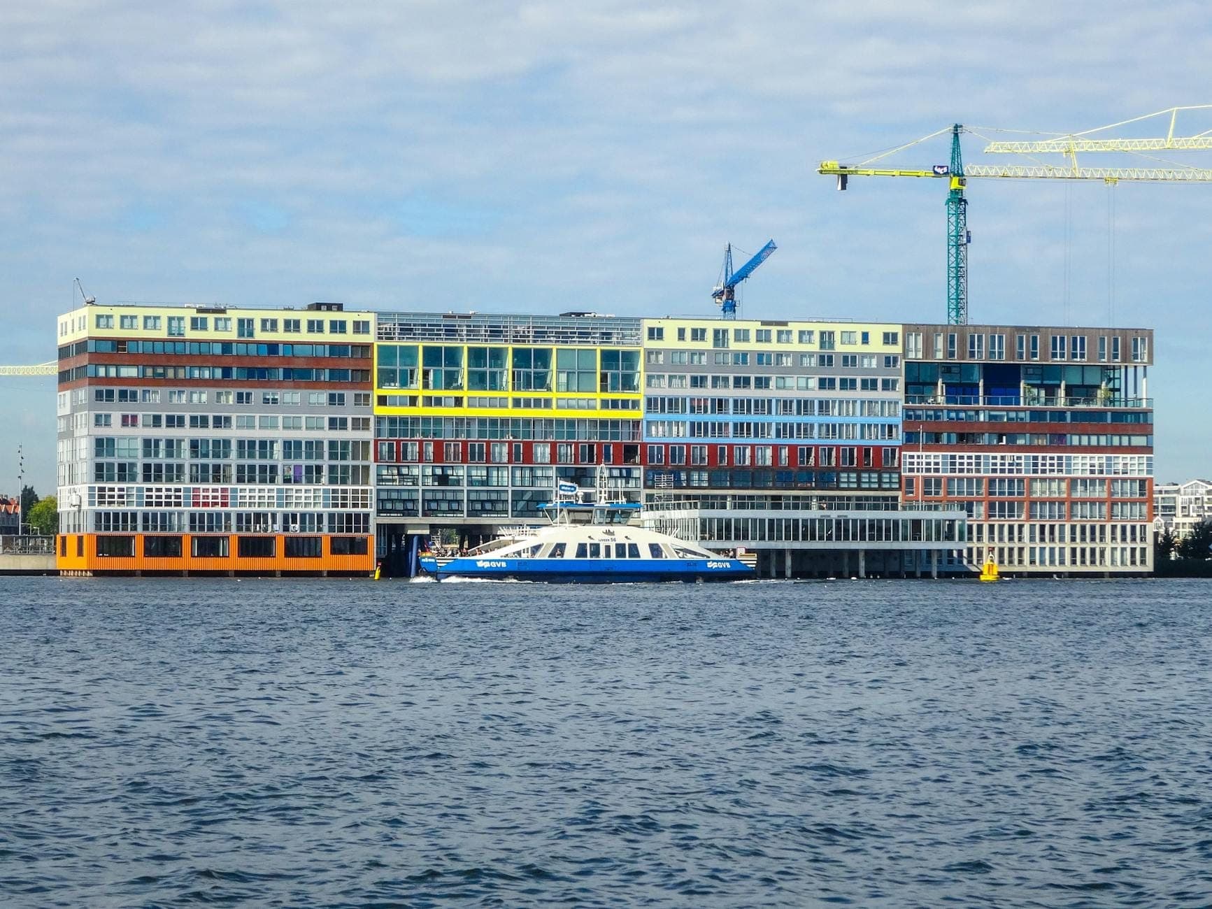 Modernes Gebäude in Amsterdam-Noord, von der anderen Seite des IJ aus gesehen, mit einer Fähre und Baukränen im Bild.