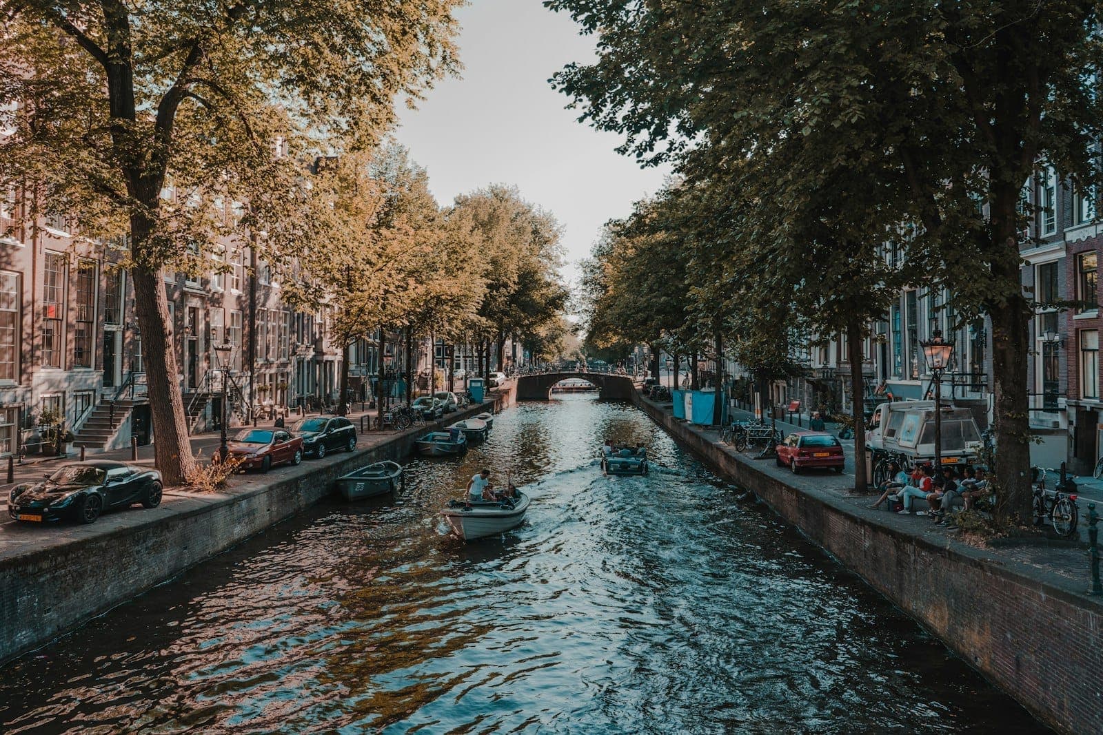 Ein ruhiger Amsterdamer Kanal, eingerahmt von Laubbäumen und traditionellen Grachtenhäusern, mit kleinen Booten auf dem Wasser und Menschen, die sich bei sanftem Tageslicht am Ufer entspannen.