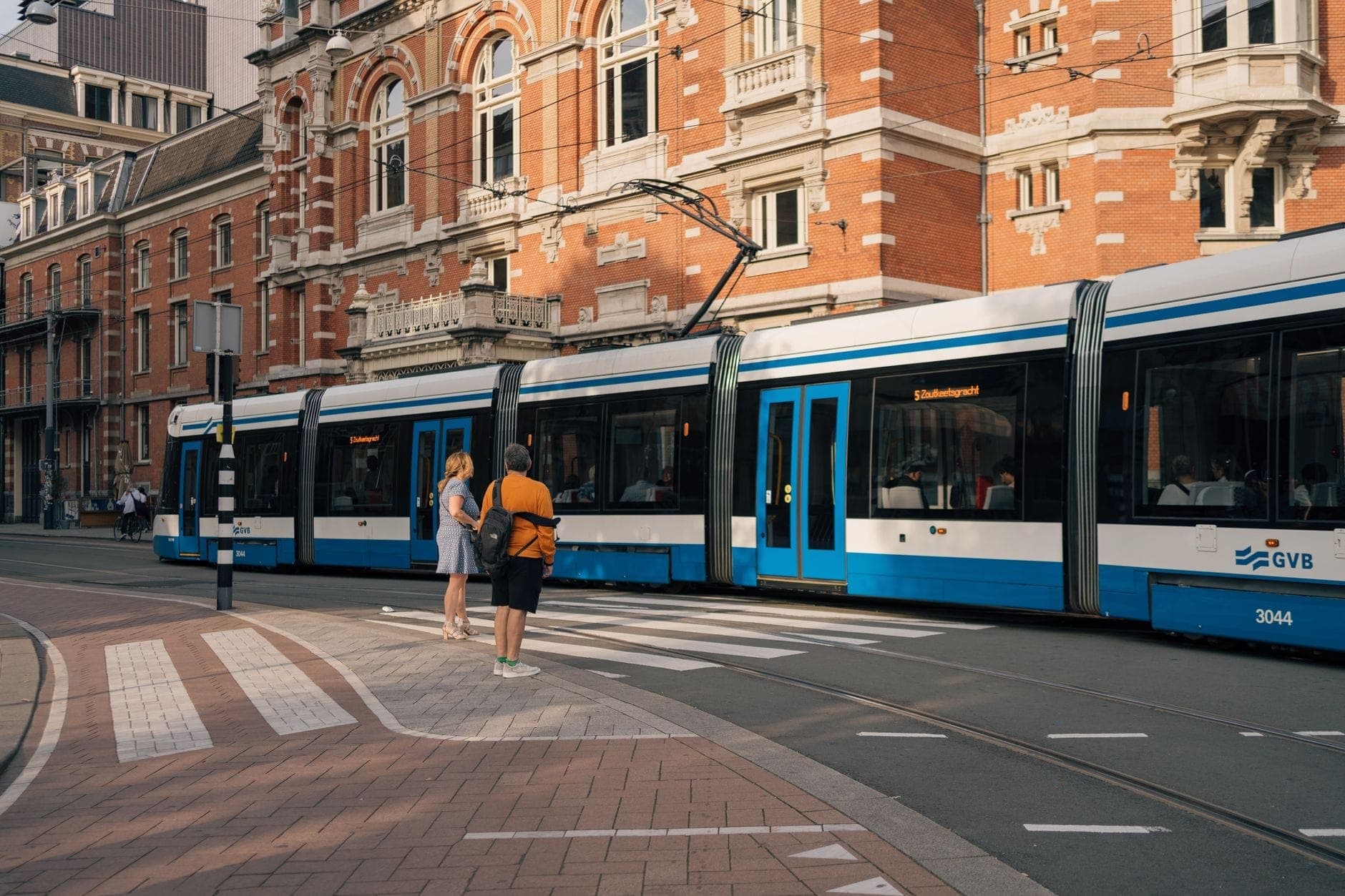 Moderne blau-weiße GVB-Straßenbahn mit wartenden Personen an einer Haltestelle vor traditioneller Amsterdamer Architektur.