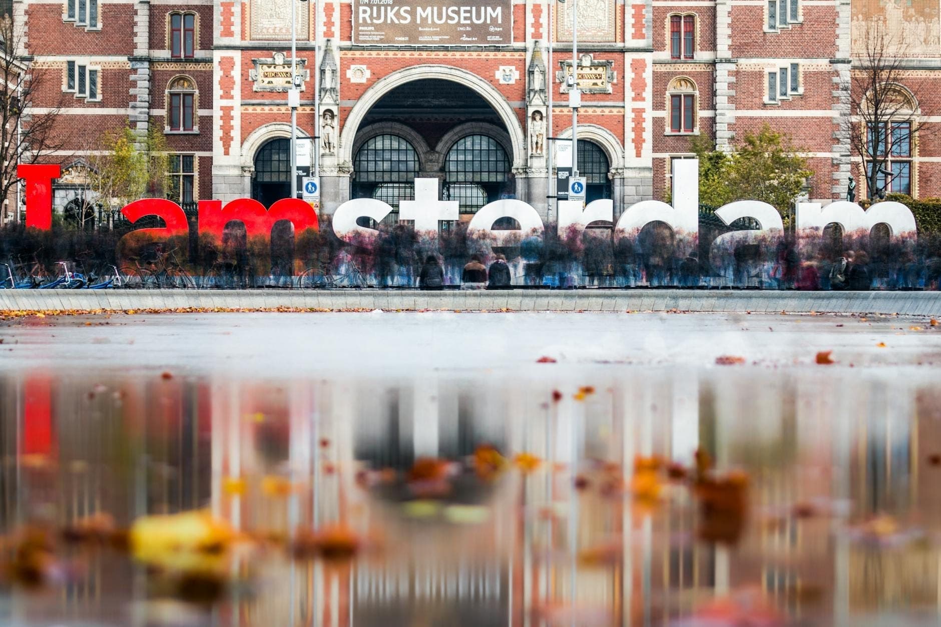 Blick auf das Rijksmuseum mit dem berühmten 'I amsterdam'-Schriftzug und Herbstblättern, die im Spiegelbecken davor treiben.