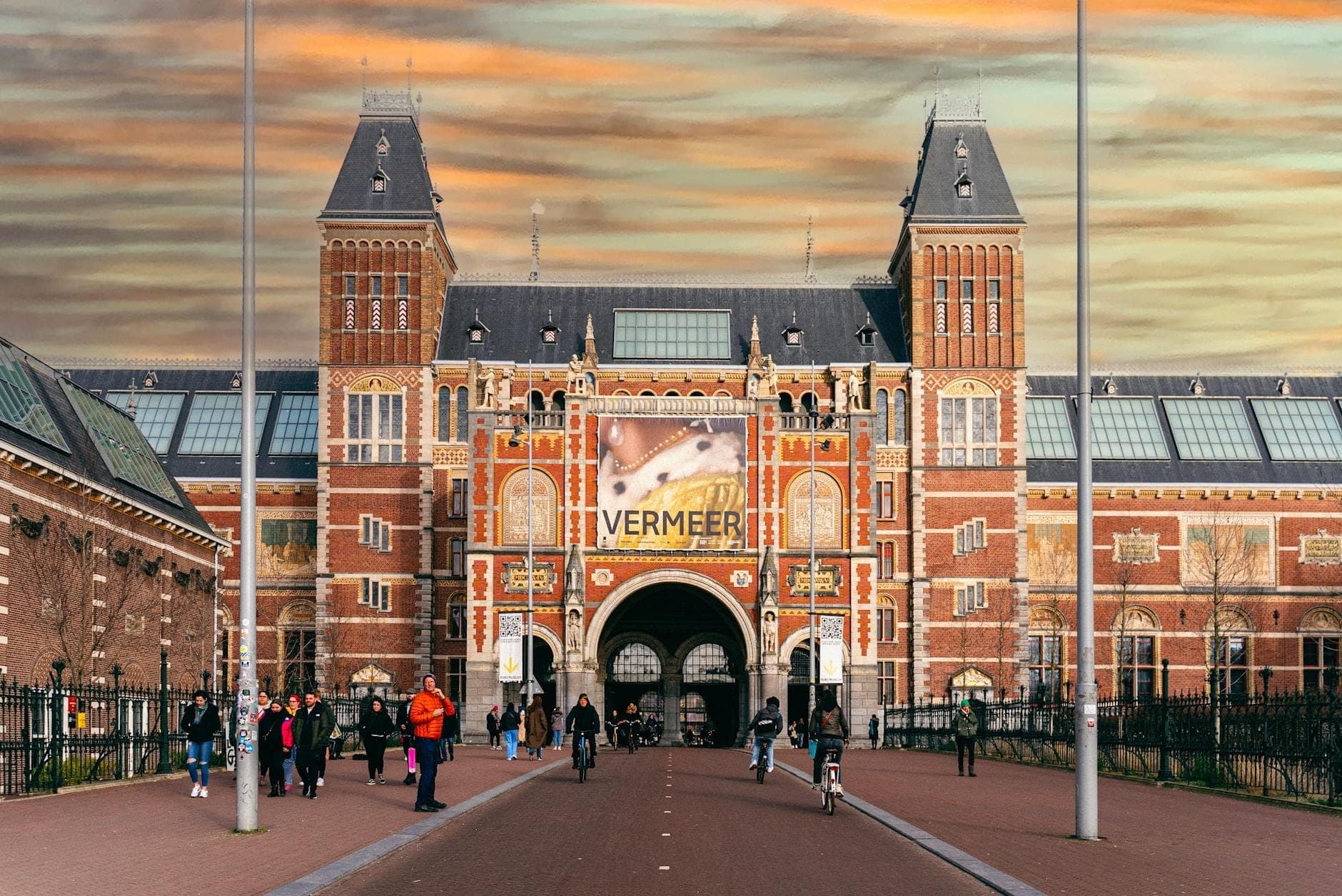 Vorderansicht des Rijksmuseums in Amsterdam mit Menschen, die unter einem bewölkten Winterhimmel spazieren und Fahrrad fahren. Vermeer-Banner ausgestellt.