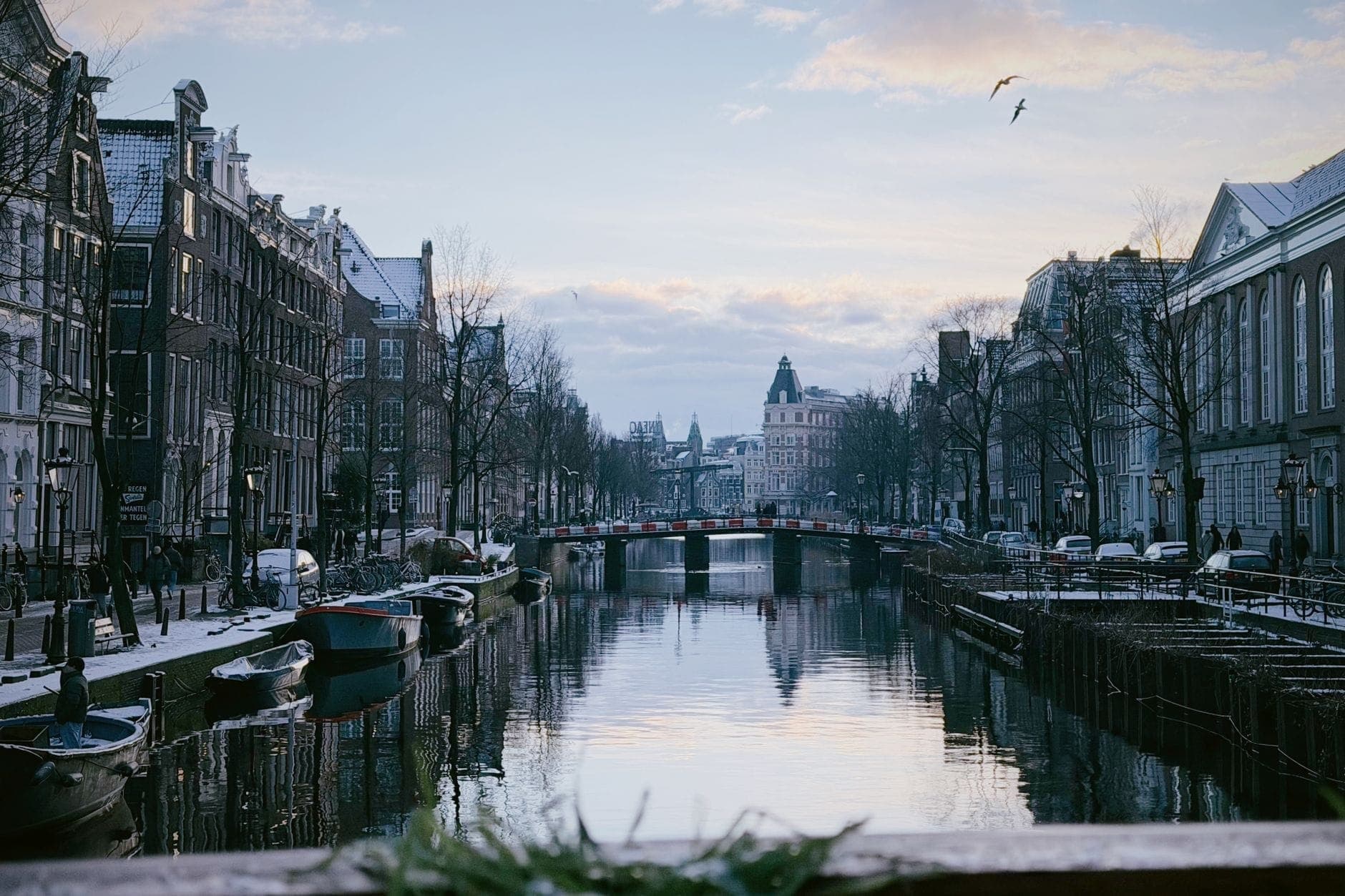 Winterszene in Amsterdam mit kahlen Bäumen, historischen Grachtenhäusern, Booten und einer Brücke in der Abenddämmerung, mit etwas Schnee auf den Dächern.