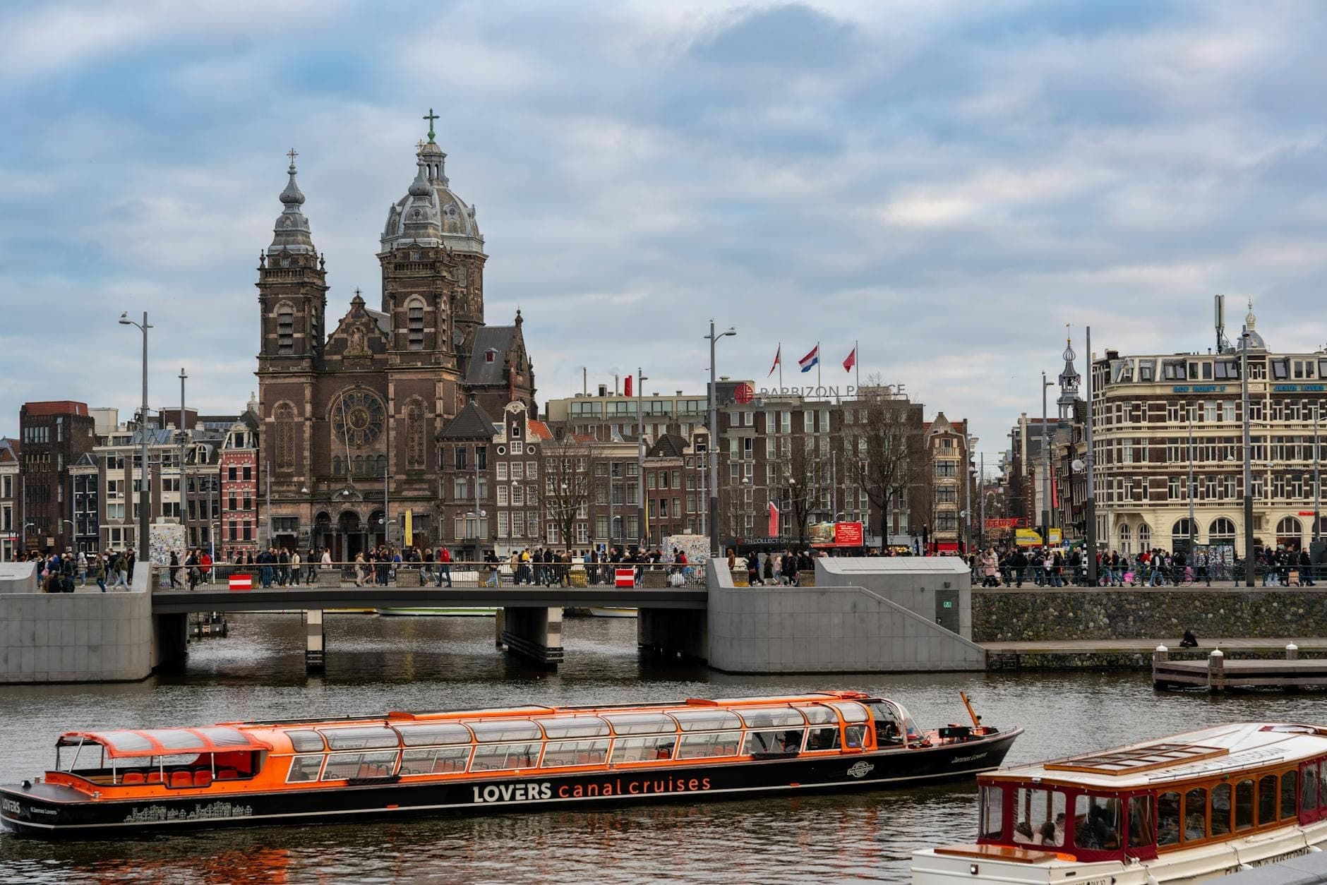 Kanalkreuzfahrtboote im Zentrum von Amsterdam mit der historischen Basilika des Heiligen Nikolaus und alten Stadtgebäuden im Hintergrund.