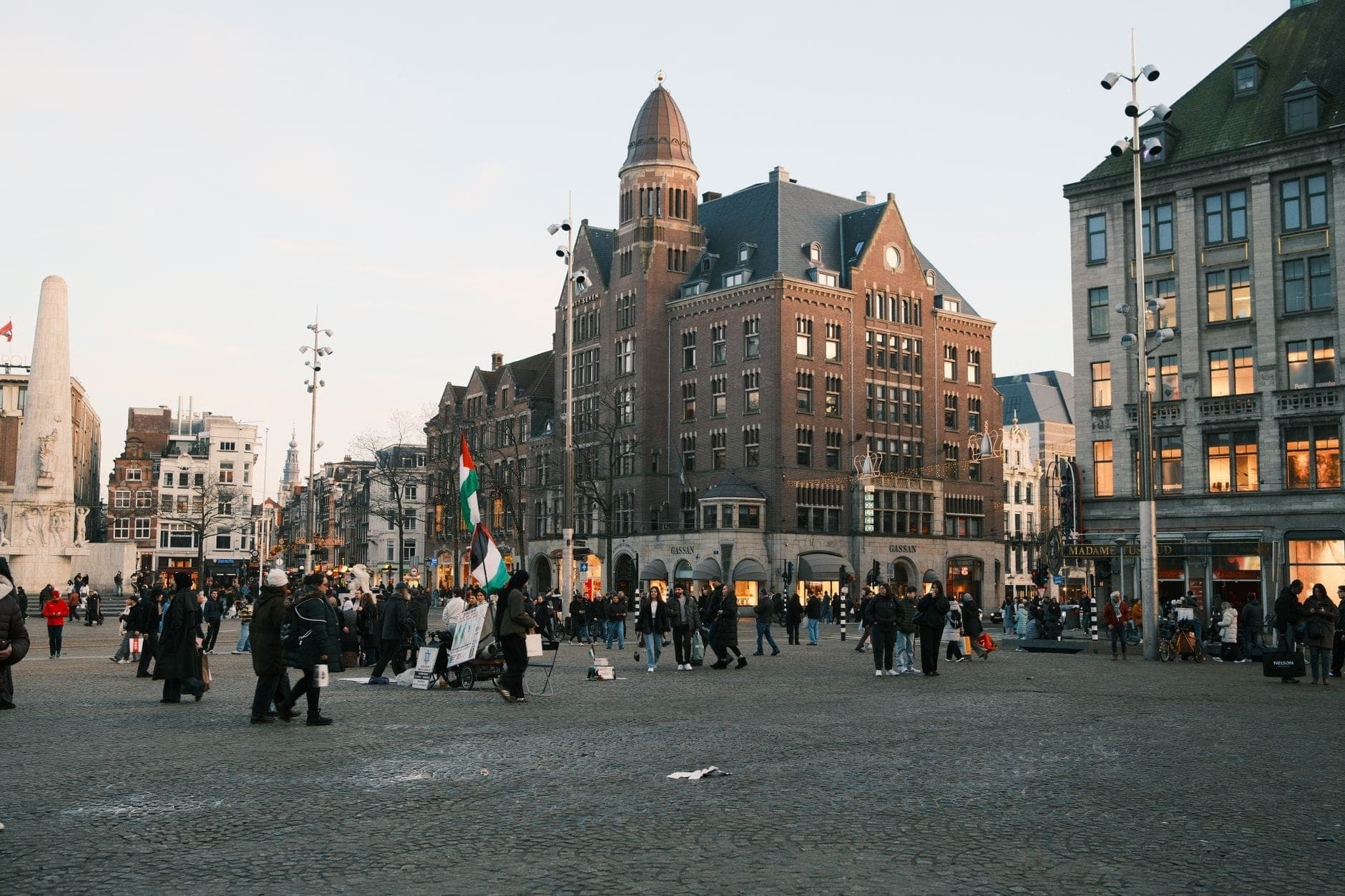 Der Dam-Platz in Amsterdam mit Spaziergängern und historischen Gebäuden, darunter der markante spitze Turm des NH Grand Hotel Krasnapolsky.