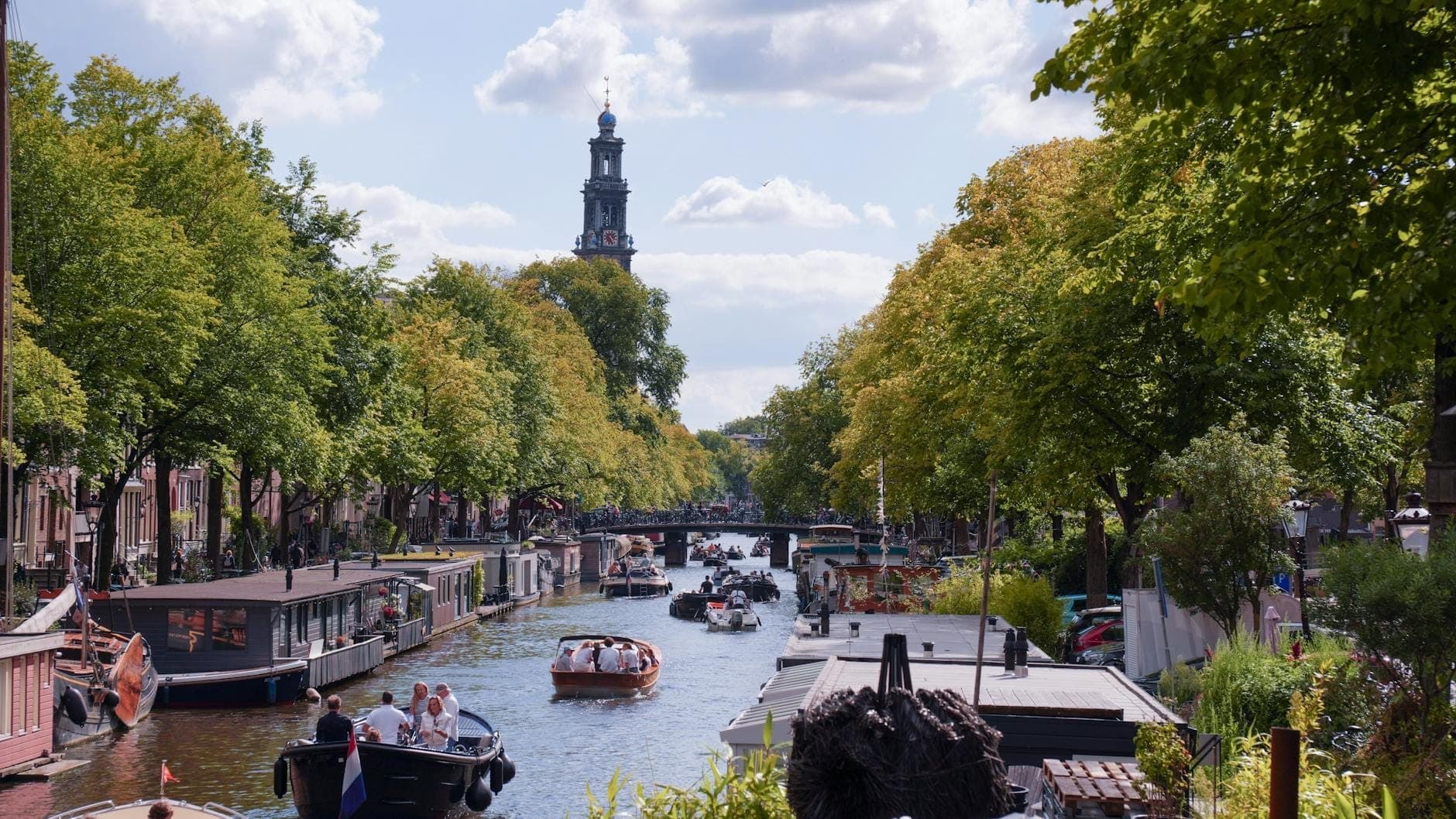 Blick auf einen Amsterdamer Kanal mit mehreren Booten, belaubten Bäumen, Hausbooten und dem Turm der Westerkerk im Hintergrund unter blauem Himmel.